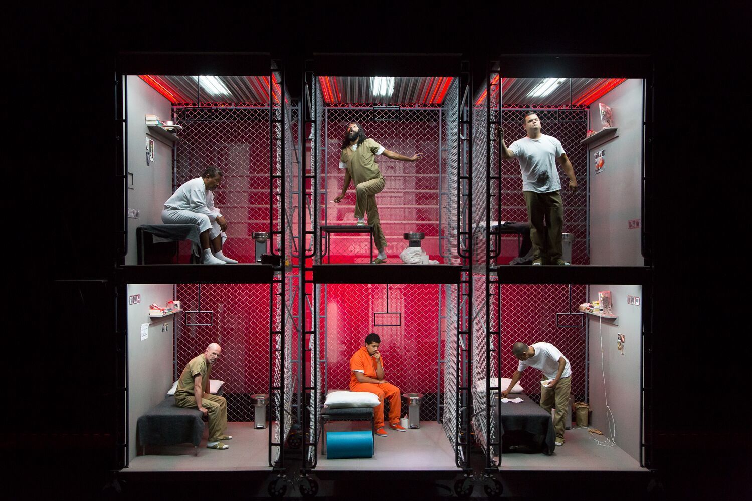 A photo of six actors on a stage inside prison cells with a red background behind them.