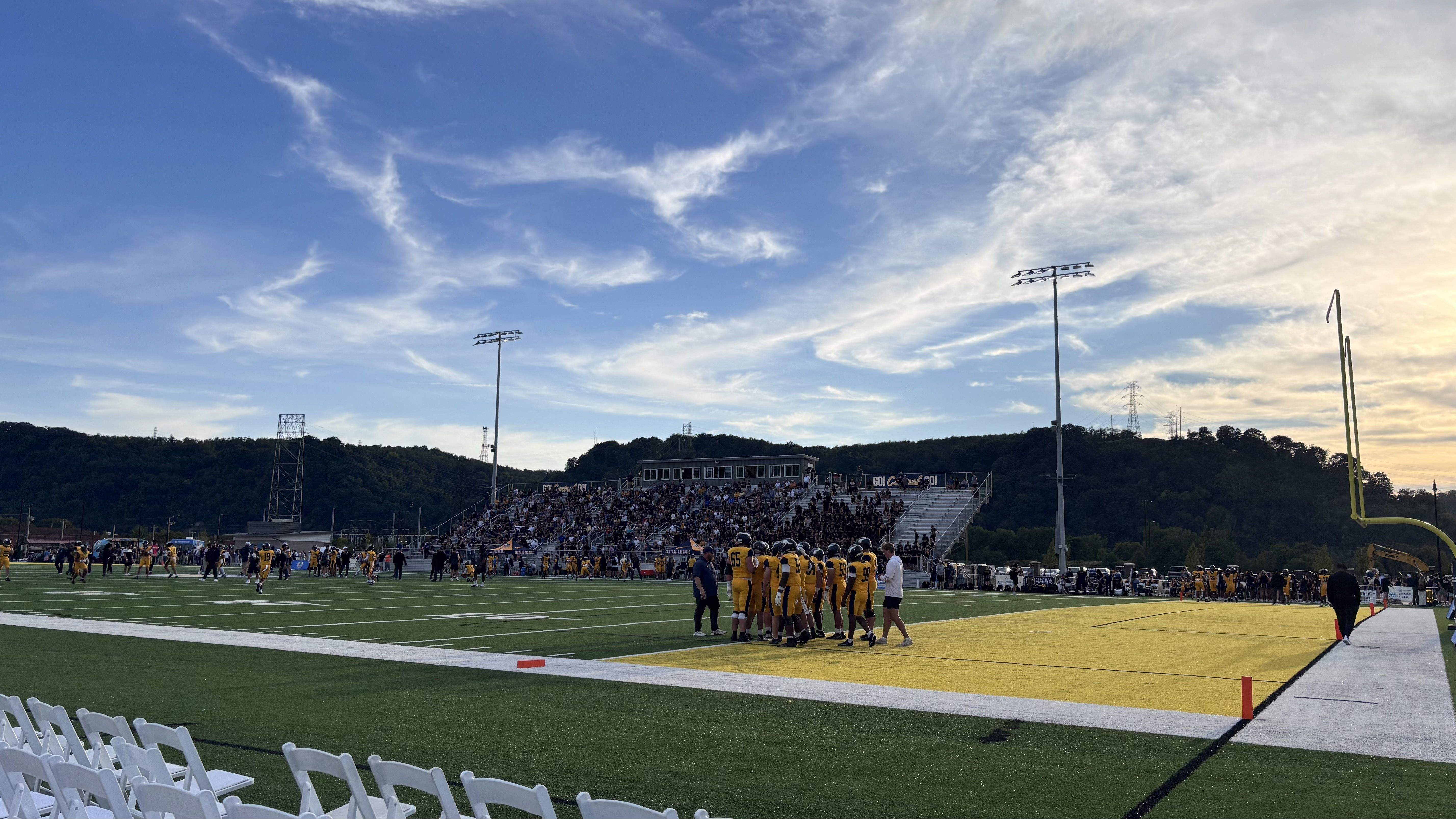 Football field on a sunny day: players in yellow near the sideline, a packed bleacher section behind, tall goalposts to the right, and a blue sky with wispy clouds over distant hills.