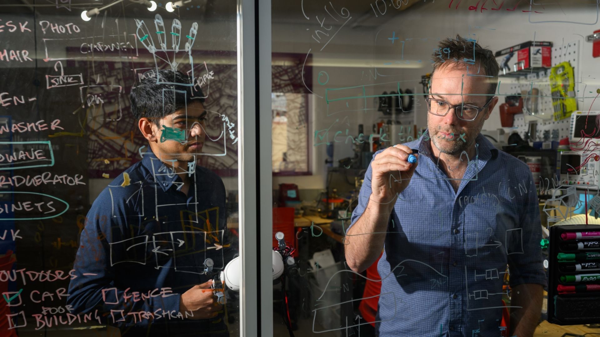 Two researchers brainstorm behind a transparent board filled with diagrams and notes in a high-tech lab setting.