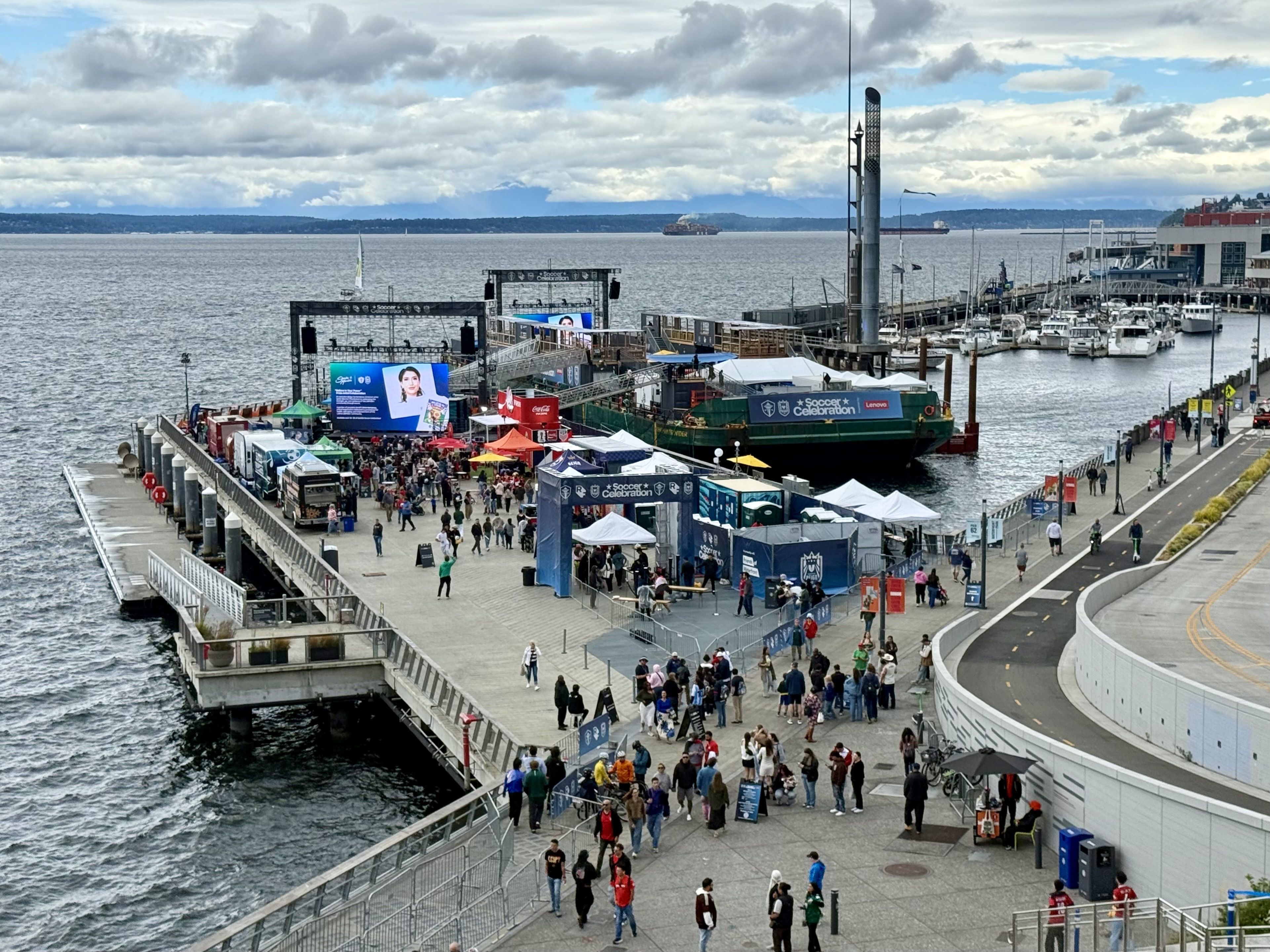 Harbor festival on a pier with a large stage and LED screens, a crowd among colorful tents, banners reading "Soccer Celebration", and boats docked at a marina under a cloudy sky.