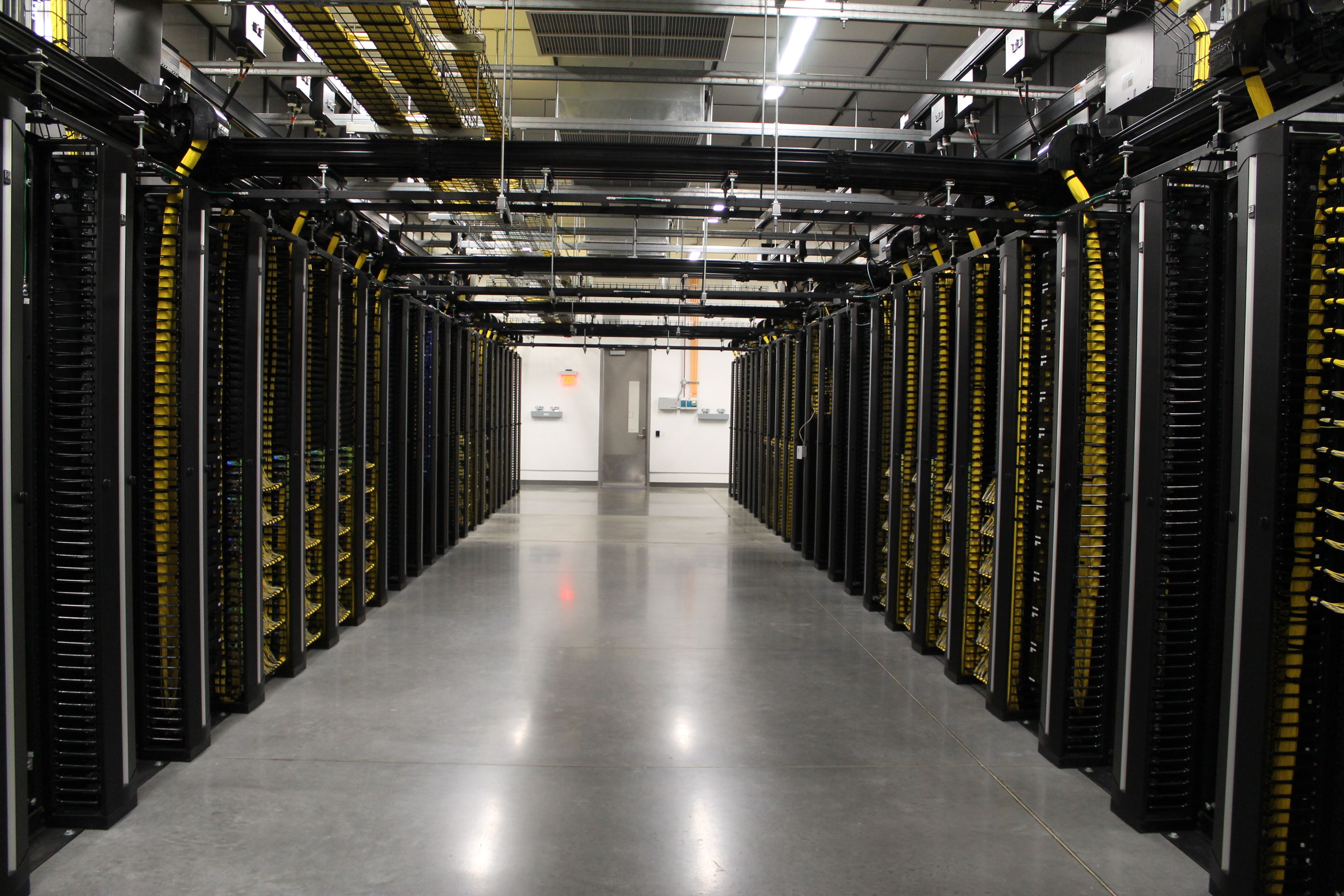 A data center aisle flanked by tall black server racks with yellow cables. Overhead cable trays and bright lights run along the ceiling; a distant door visible at end of the corridor.