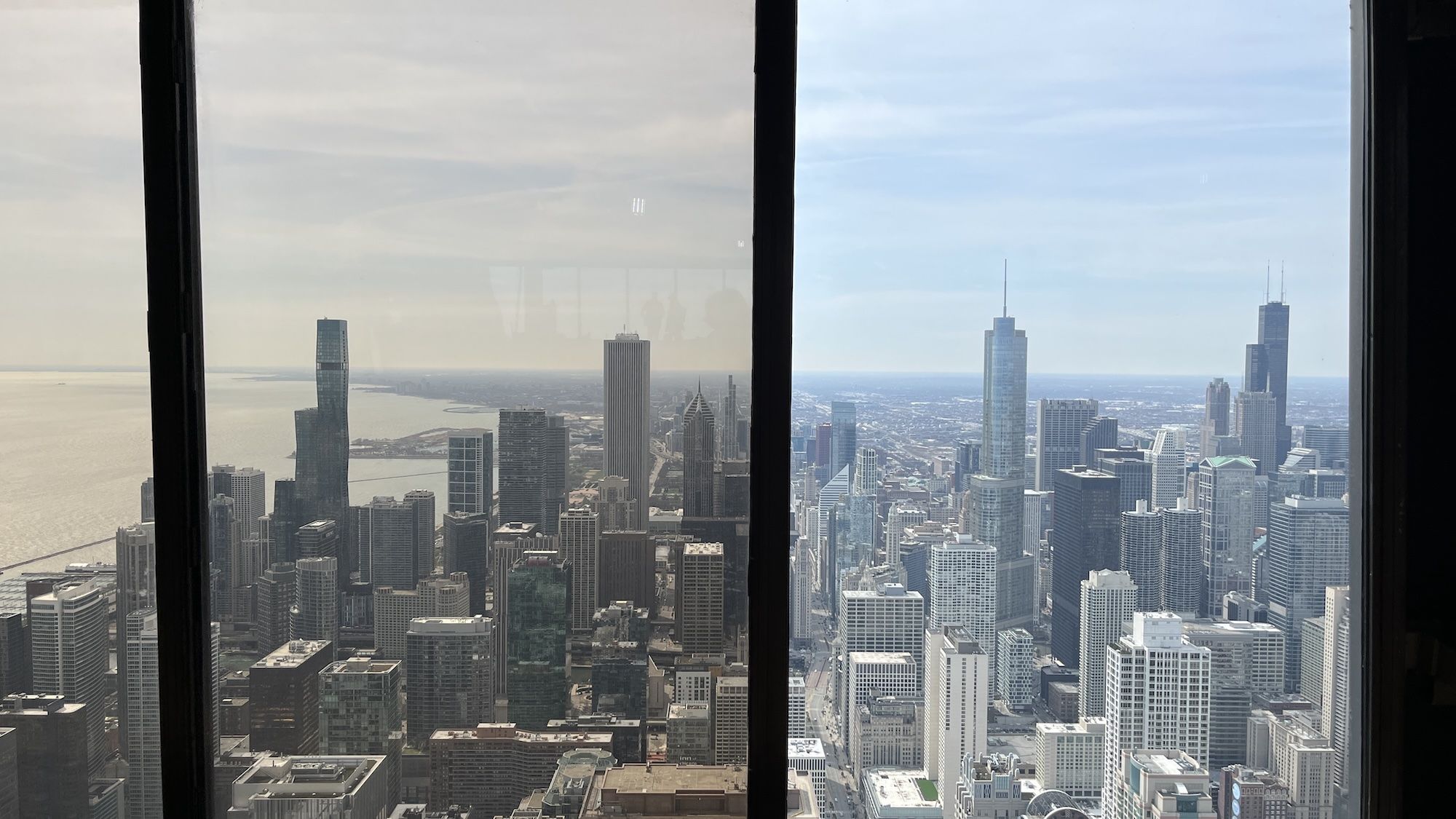 Chicago's skyline seen from the 95th floor, framed by two vertical window panes. Clusters of gray skyscrapers stretch toward the horizon beside a large body of water on the left, under a clear blue sky.