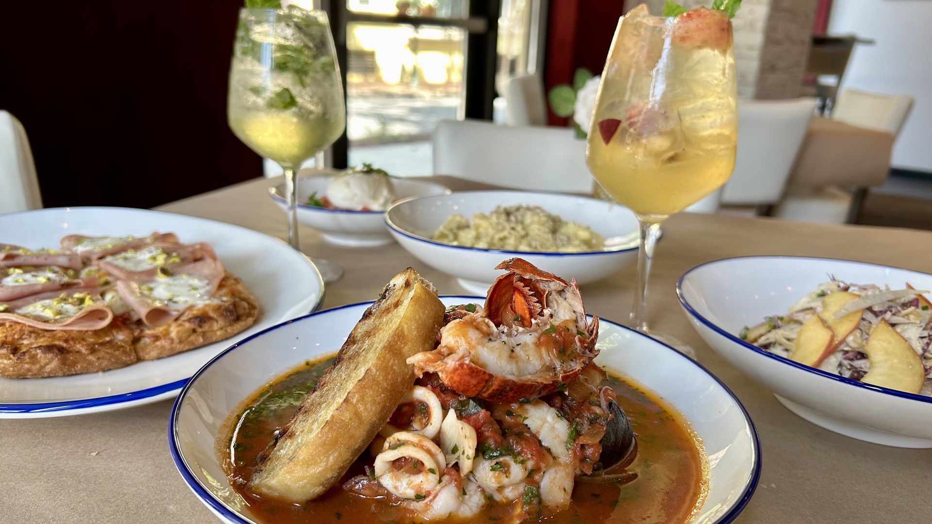 Table with seafood stew including shrimp and lobster with toasted bread, a ham and cheese pizza, pasta, salad with sliced pears, and two yellow cocktails garnished with mint in a bright restaurant.