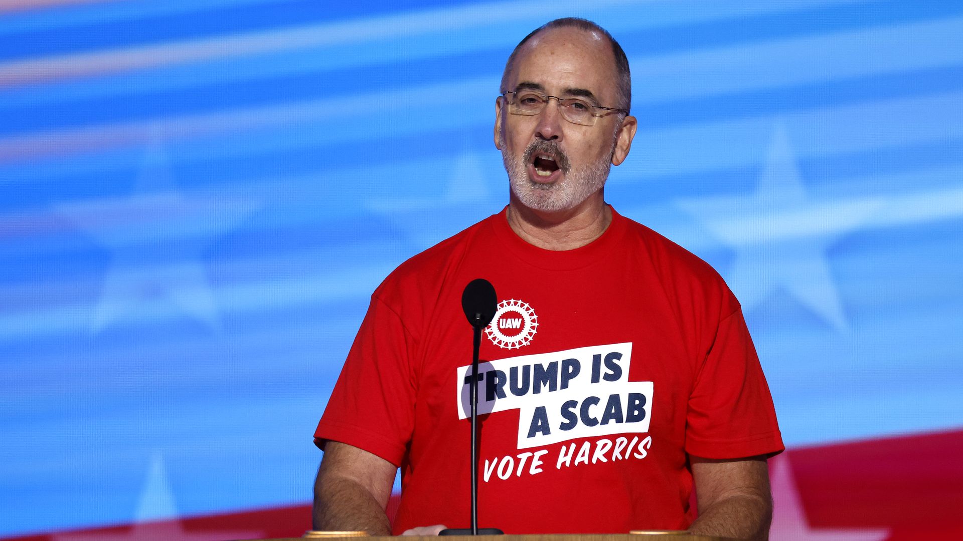 A person in a red T-shirt stating "TRUMP IS A SCAB; VOTE HARRIS" speaks at a lectern 