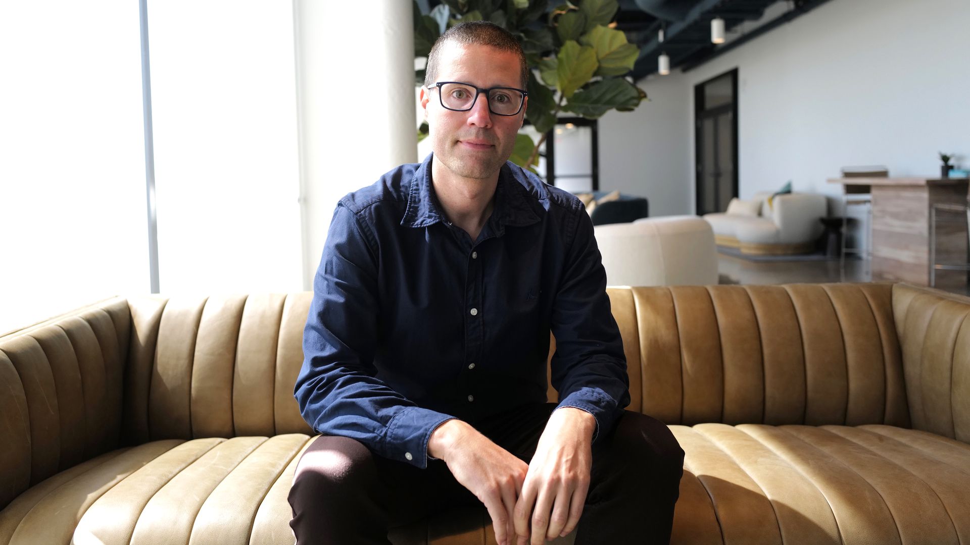 A photo of a man sitting in the middle of a leather couch in an office building hallway.