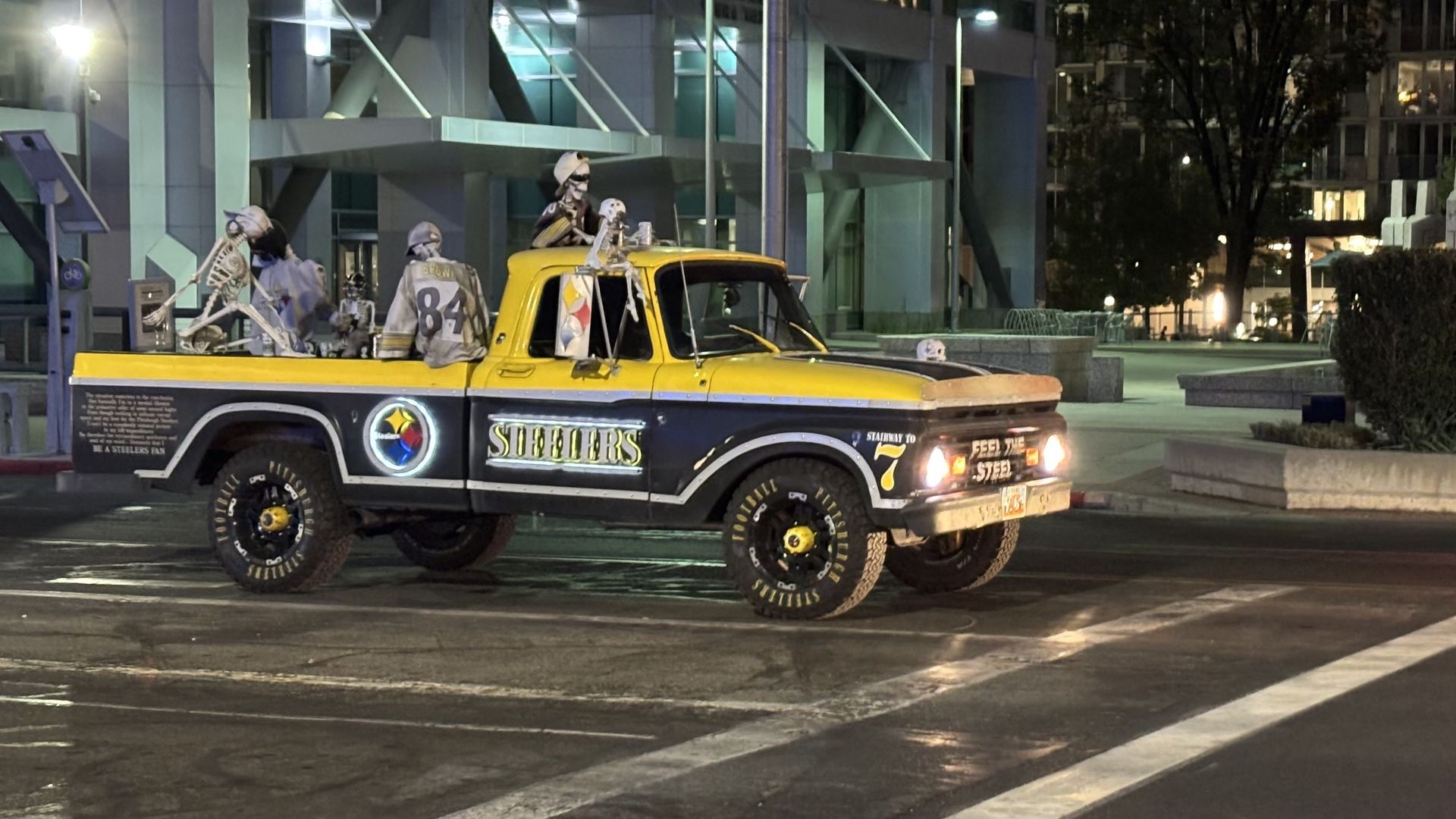 Yellow and black Steelers-themed pickup truck at night with skeleton decorations wearing helmets and jerseys, parked near modern buildings and lit streetlamps.