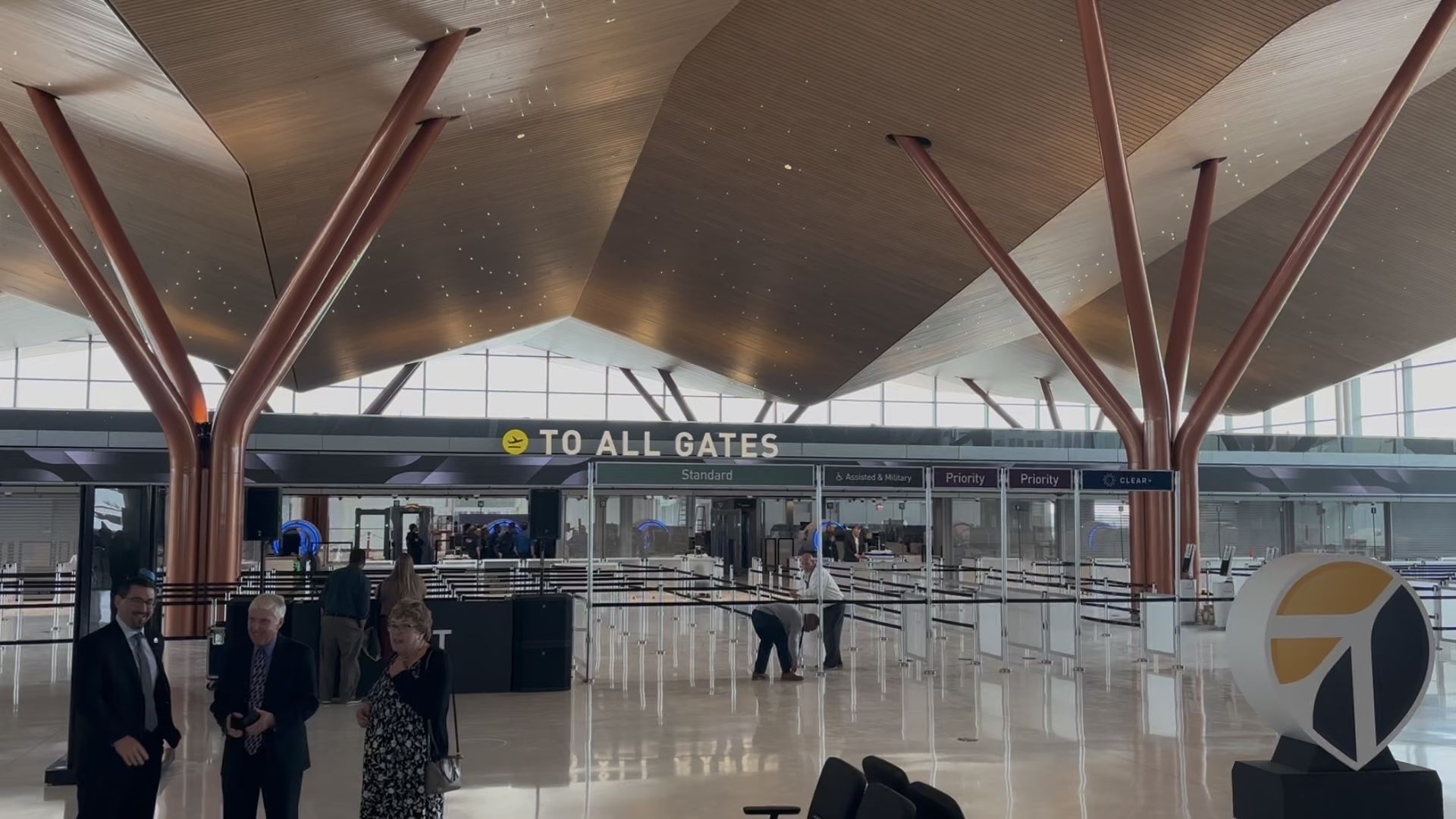 Airport security checkpoint with signs for Standard, Assisted & Military, and Priority lanes under large wooden ceiling with red steel beams and people standing nearby.