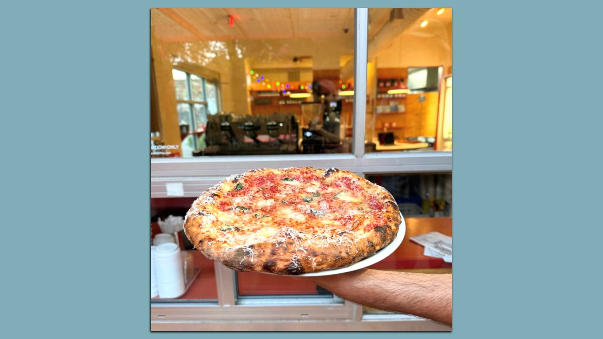 Close-up of a large cheese pizza with various toppings on a white plate, held by a person outside a shop. The restaurant interior is visible through the glass frontage with warm lights and shelves.