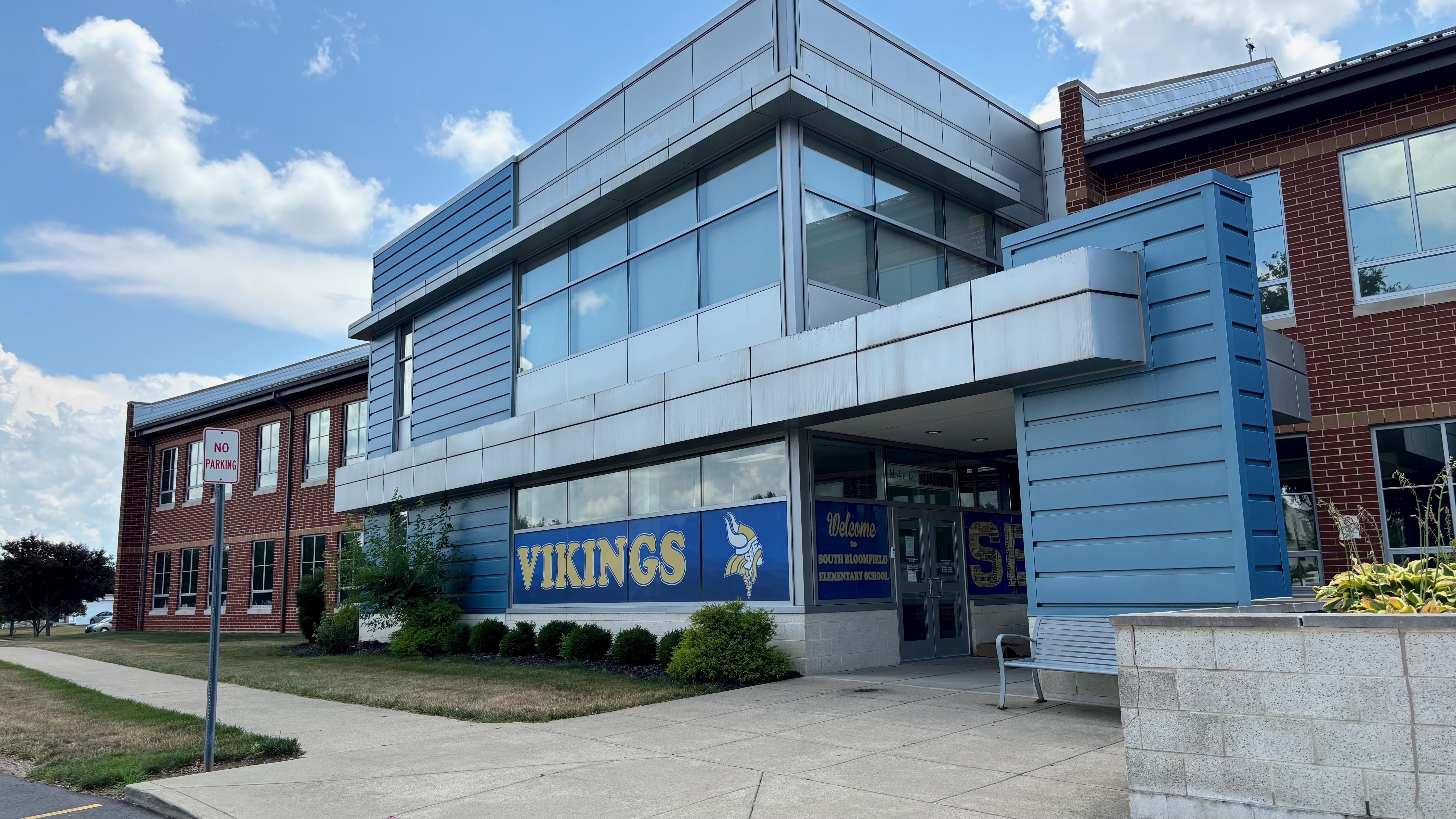 Modern school building with blue metal panels and large glass windows; the entrance displays a blue Vikings banner with a Viking mascot, brick walls, and a No Parking sign nearby.