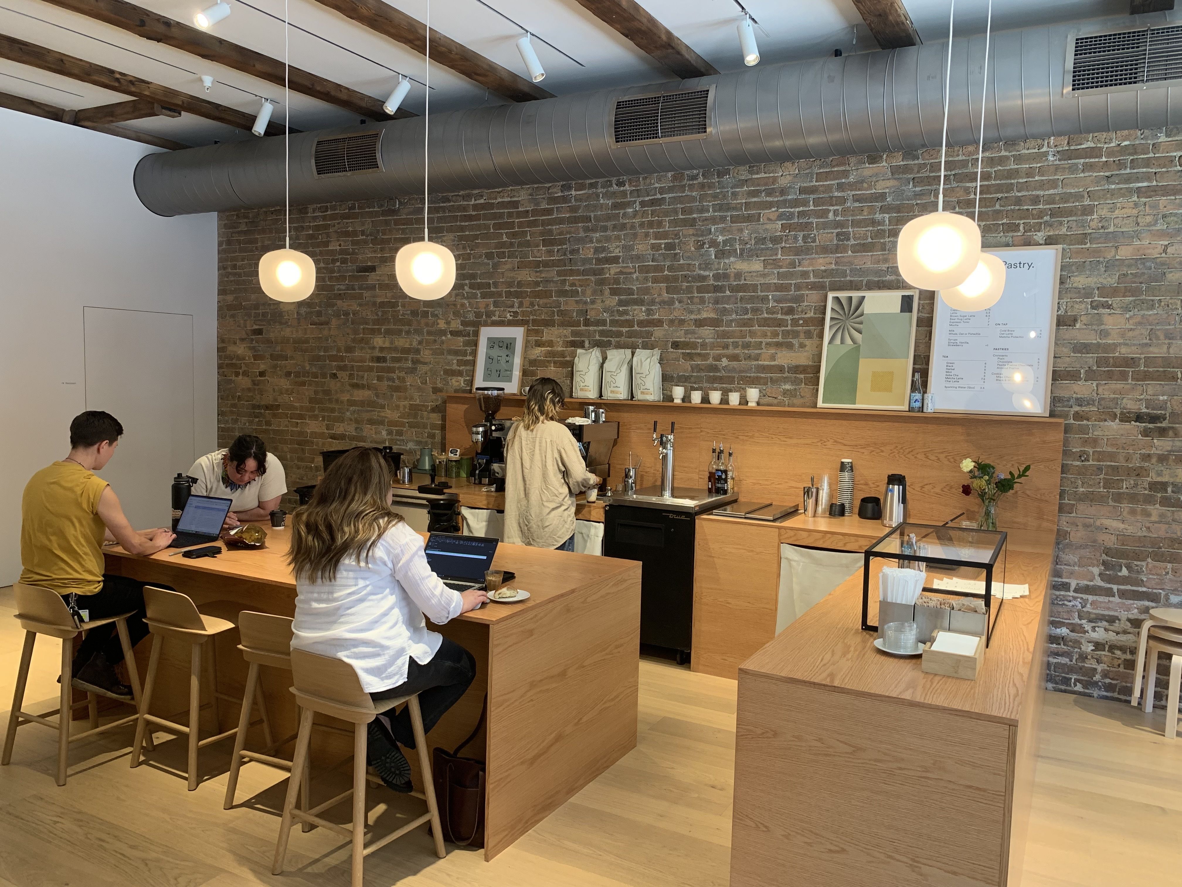 Interior of a modern cafe with an exposed brick wall, wooden ceiling beams, large grey ductwork, and round pendant lights. Customers sit at long wooden tables with laptops and coffee cups.