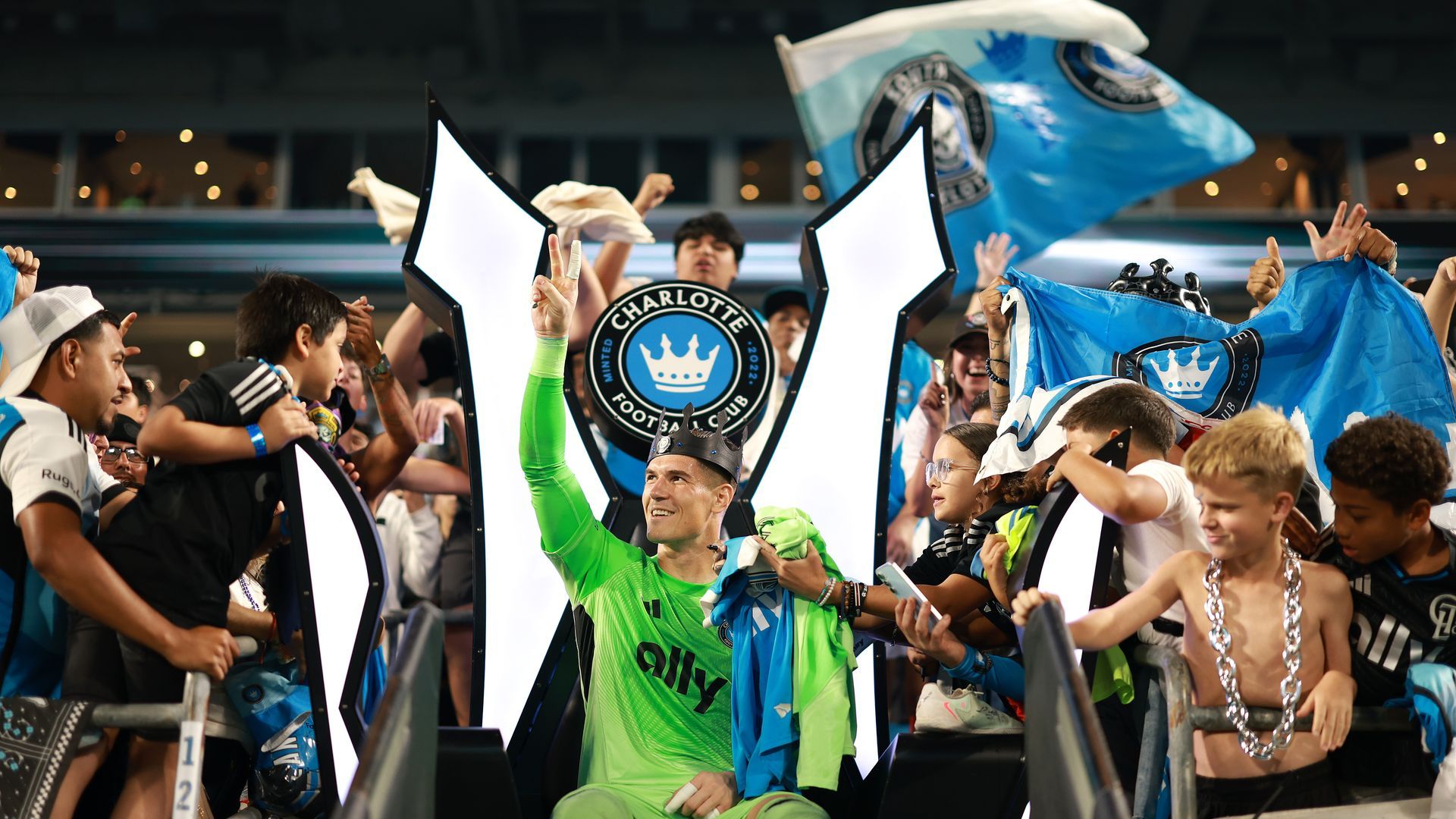 Soccer player in green goalkeeper gear and crown gestures peace sign to fans holding blue Charlotte FC flags and scarves, seated in illuminated throne-shaped chair at stadium event.