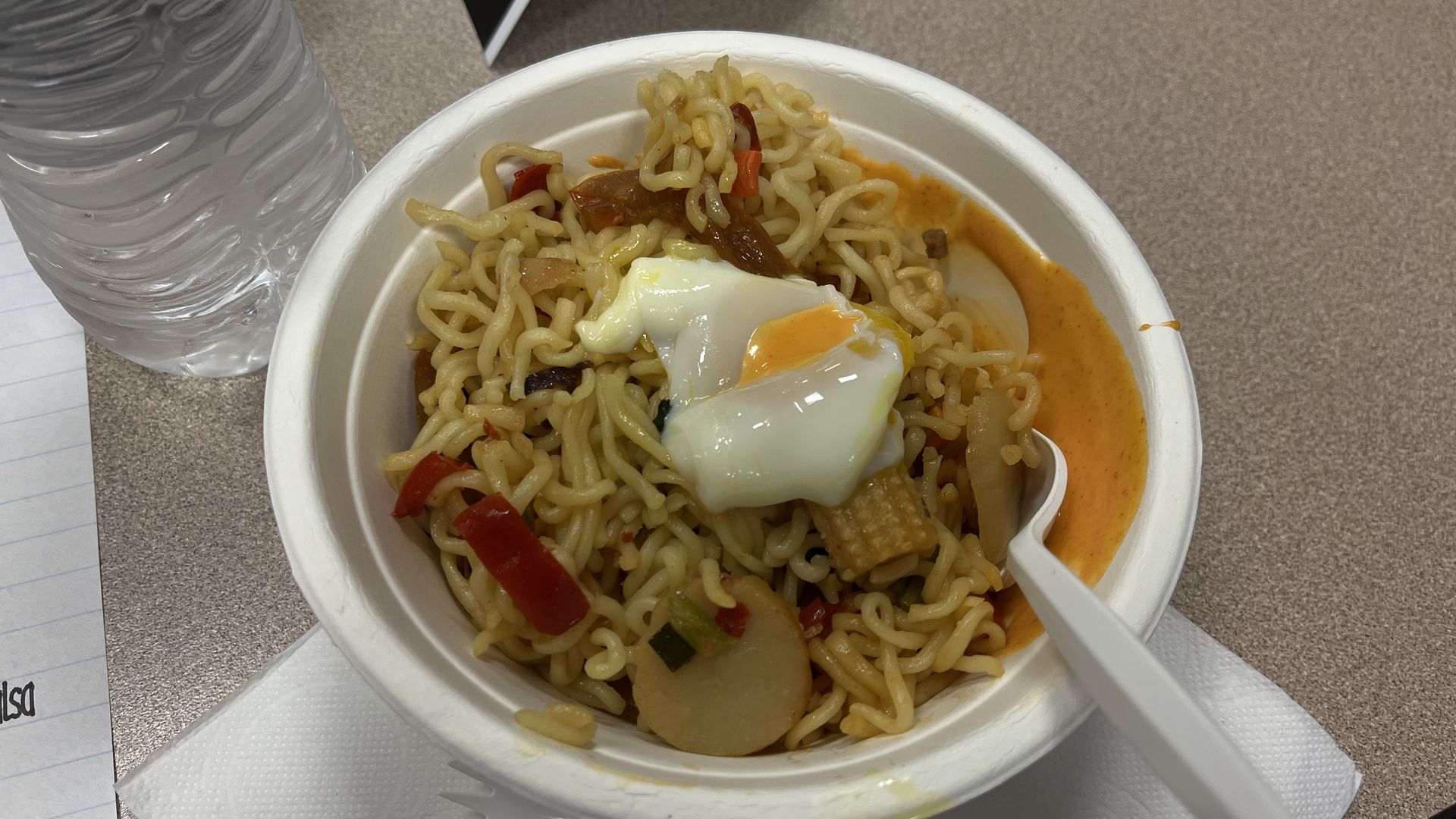White bowl with cooked ramen noodles, mixed vegetables, and a soft-boiled egg, placed on a white napkin with a plastic fork and spoon, next to a bottle of water on a beige table.