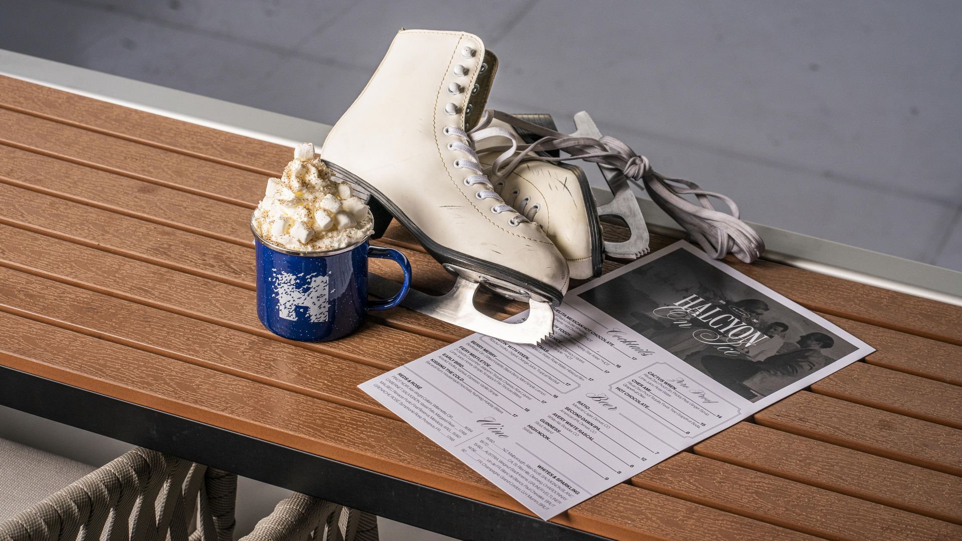 White ice skates, a blue mug topped with marshmallows and whipped cream, and a menu titled "Halcyon" placed on a wooden table with a gray background.