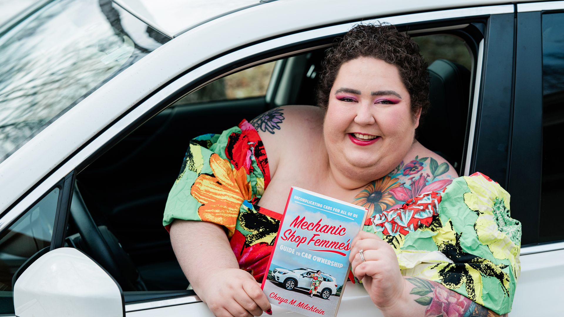 A woman in a floral dress sits in a car, holding a copy of the  new "Mechanic Shop Femme's" book. 