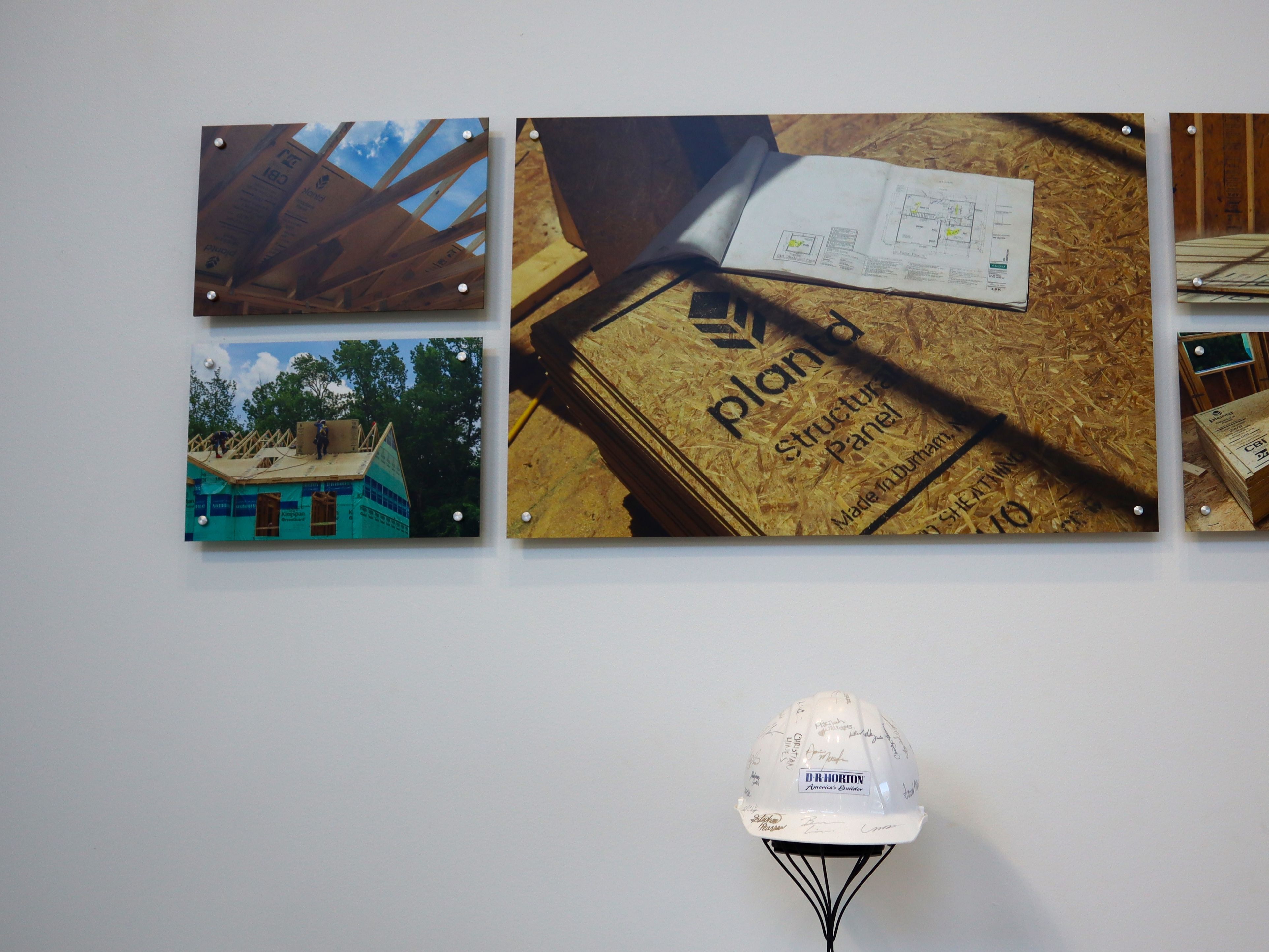 Construction photo collage on a white wall: left panels show framing and a partially built house; center panel shows plywood with a plan and an open notebook; a signed white safety helmet sits on a stand below.
