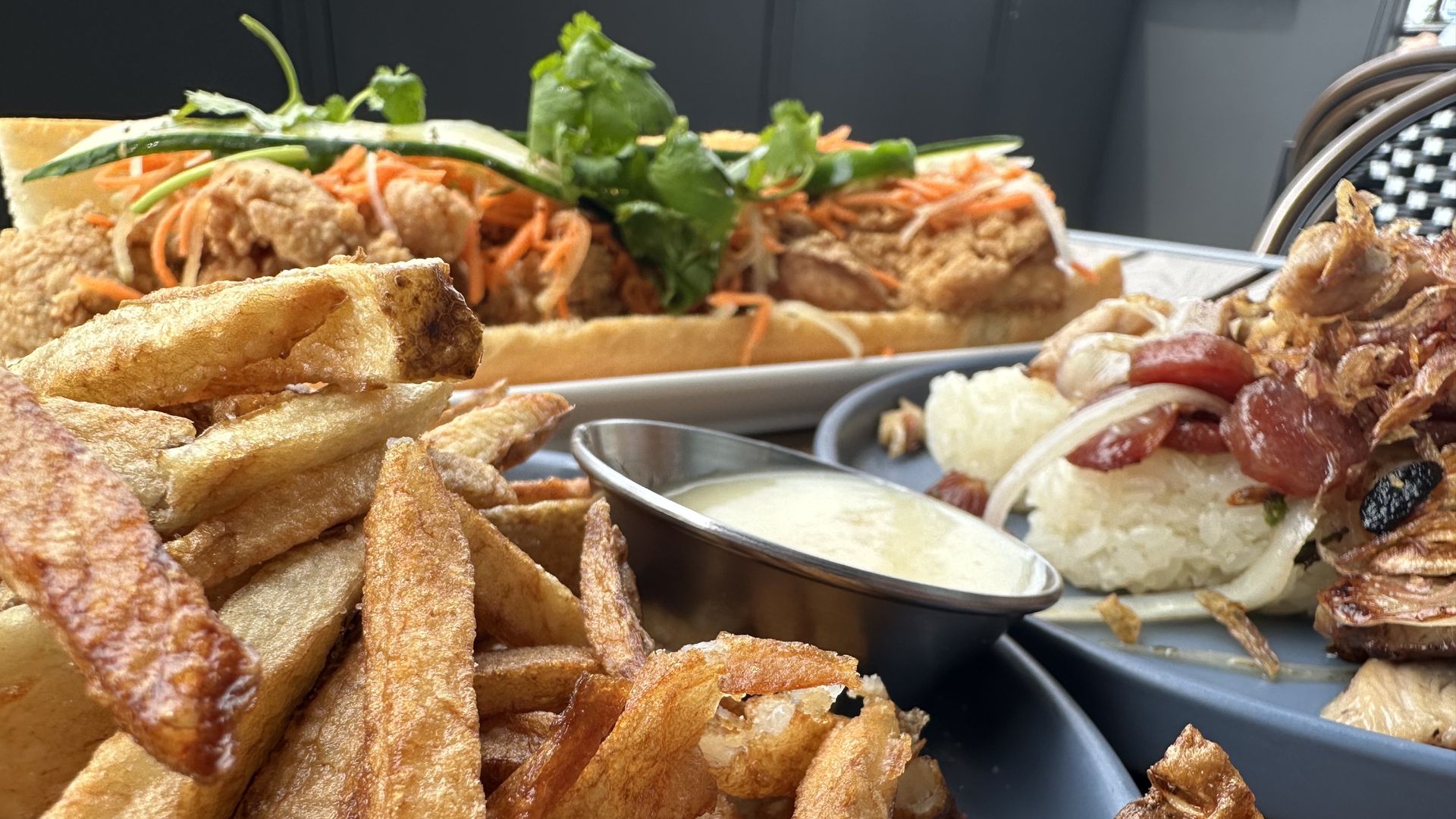 A fried chicken banh mi, a plate of sticky rice and french fries with an aioli sauce sit on a table outside.