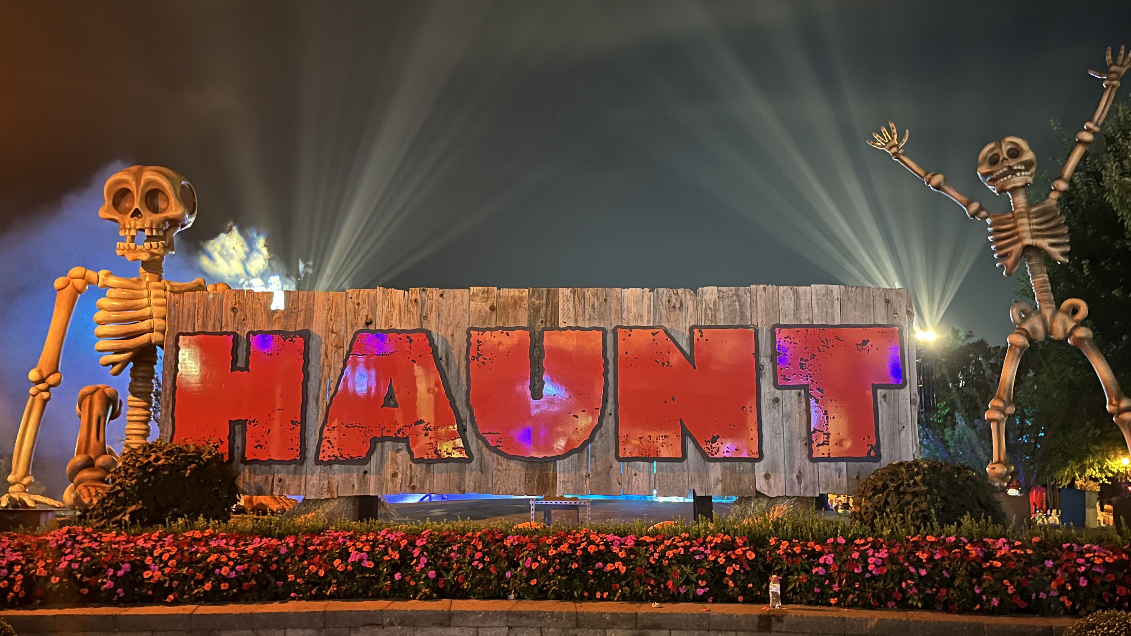 Night scene with large wooden sign reading HAUNT in red letters, flanked by two tall, orange skeleton figures, bright spotlights in background, and colorful flowerbed in the foreground.