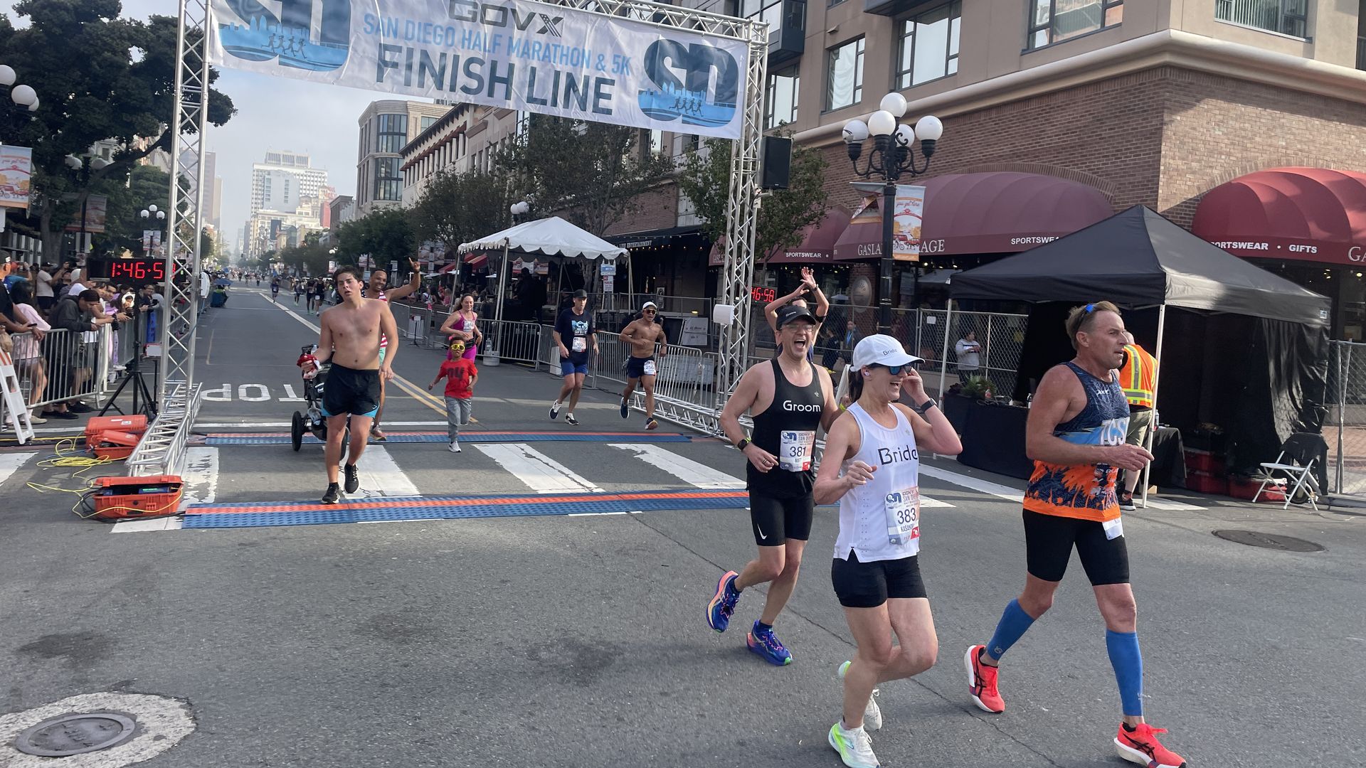 Runners cross the finish line at the San Diego Half Marathon under a banner; spectators cheer behind barriers on a city street. In the foreground, a shirtless man and a couple wear "Bride" and "Groom" shirts.