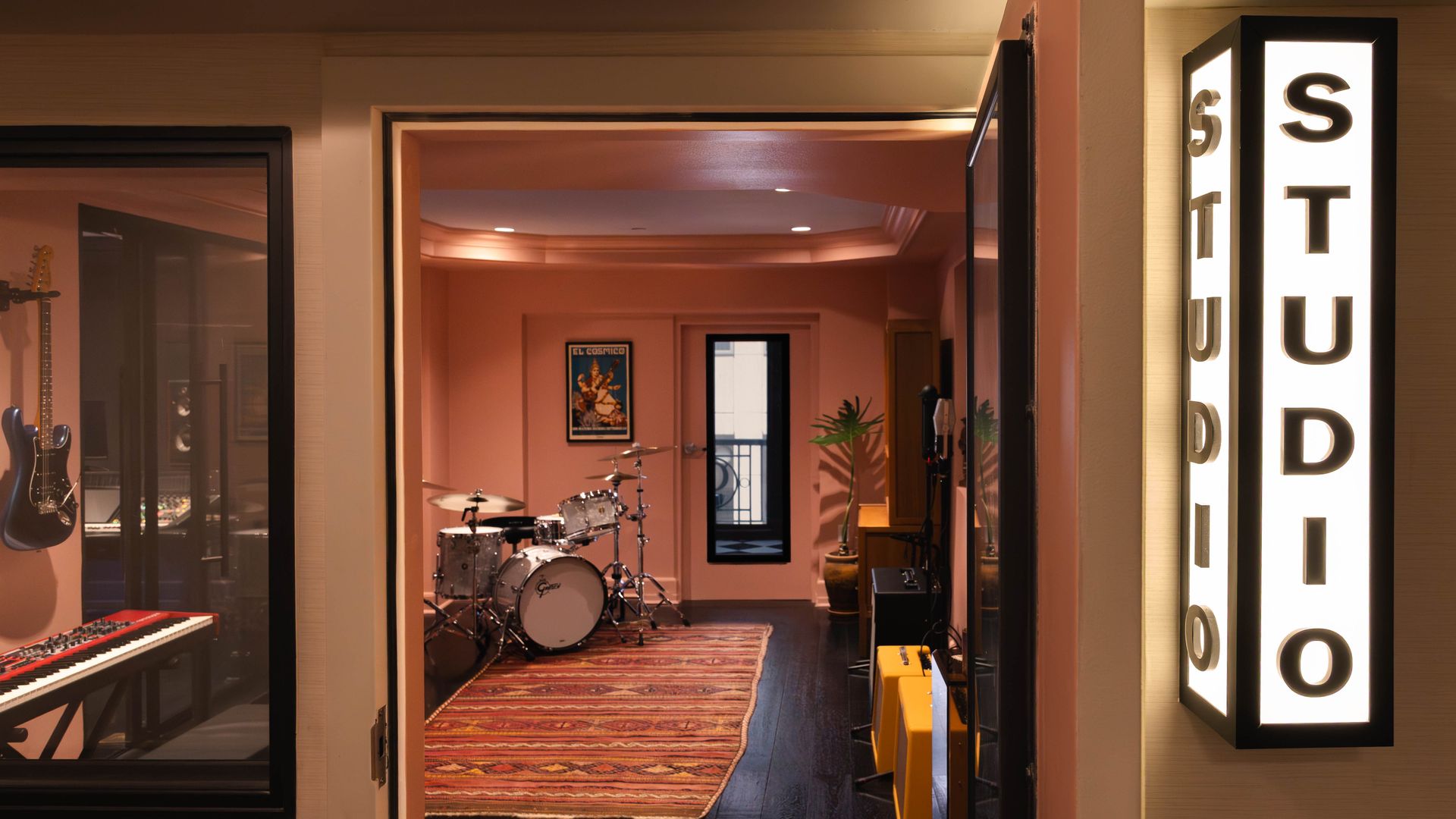 Music studio hallway with a drum kit visible in a pink-walled inner room, glass-walled guitar area to the left, a red keyboard front left, and a tall illuminated STUDIO sign on the right.