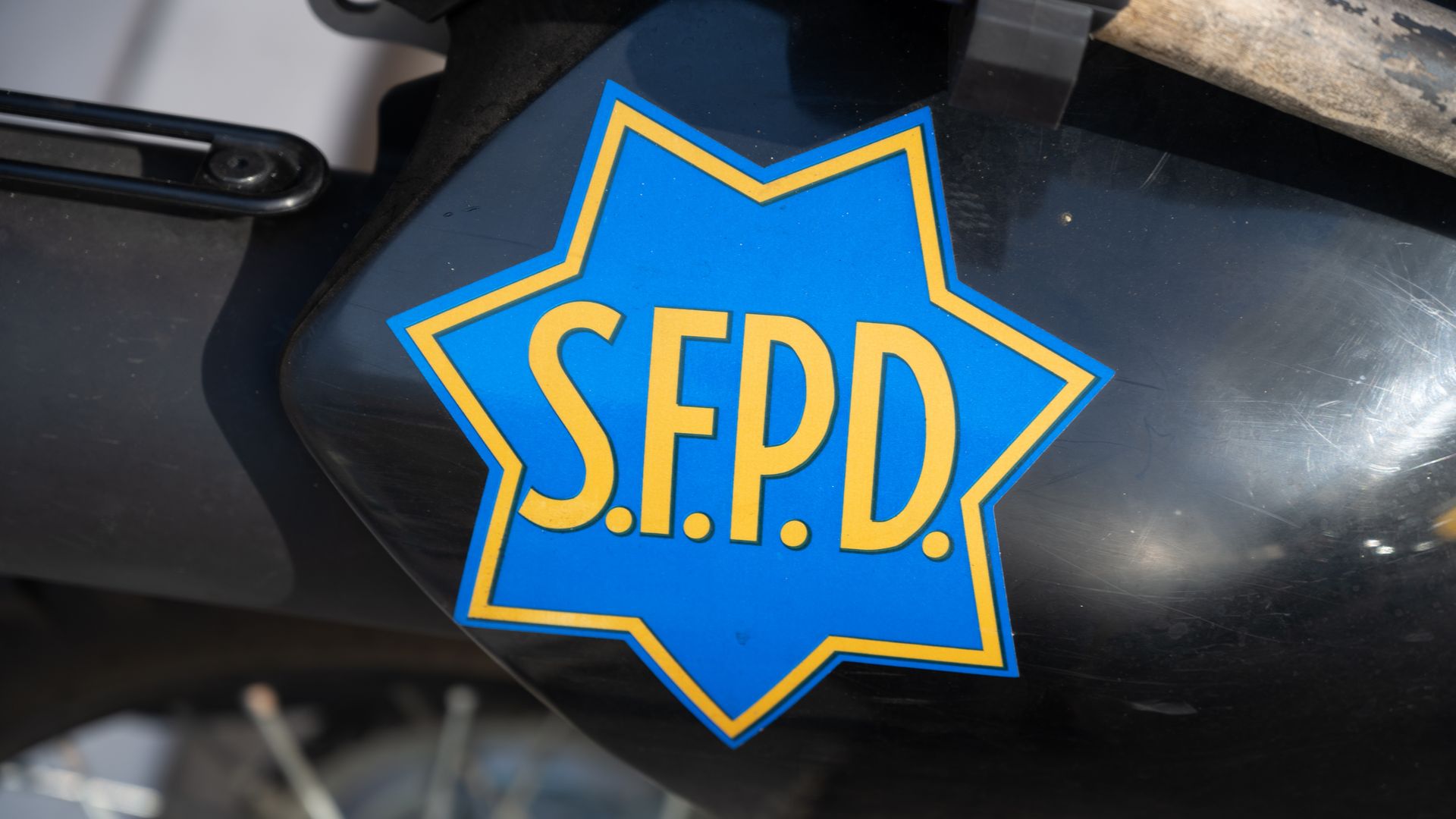 Blue eight-point star police badge with a yellow border and the letters S.F.P.D., resting on a dark surface of a police motorcycle with chrome accents in the background.