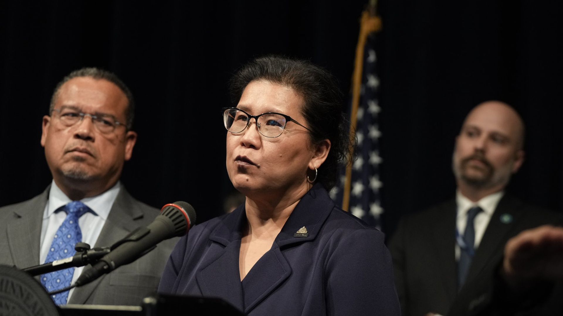 Mayor Kaohly Her stands in a navy suit at a microphone with Attorney General Keith Ellison looking on.