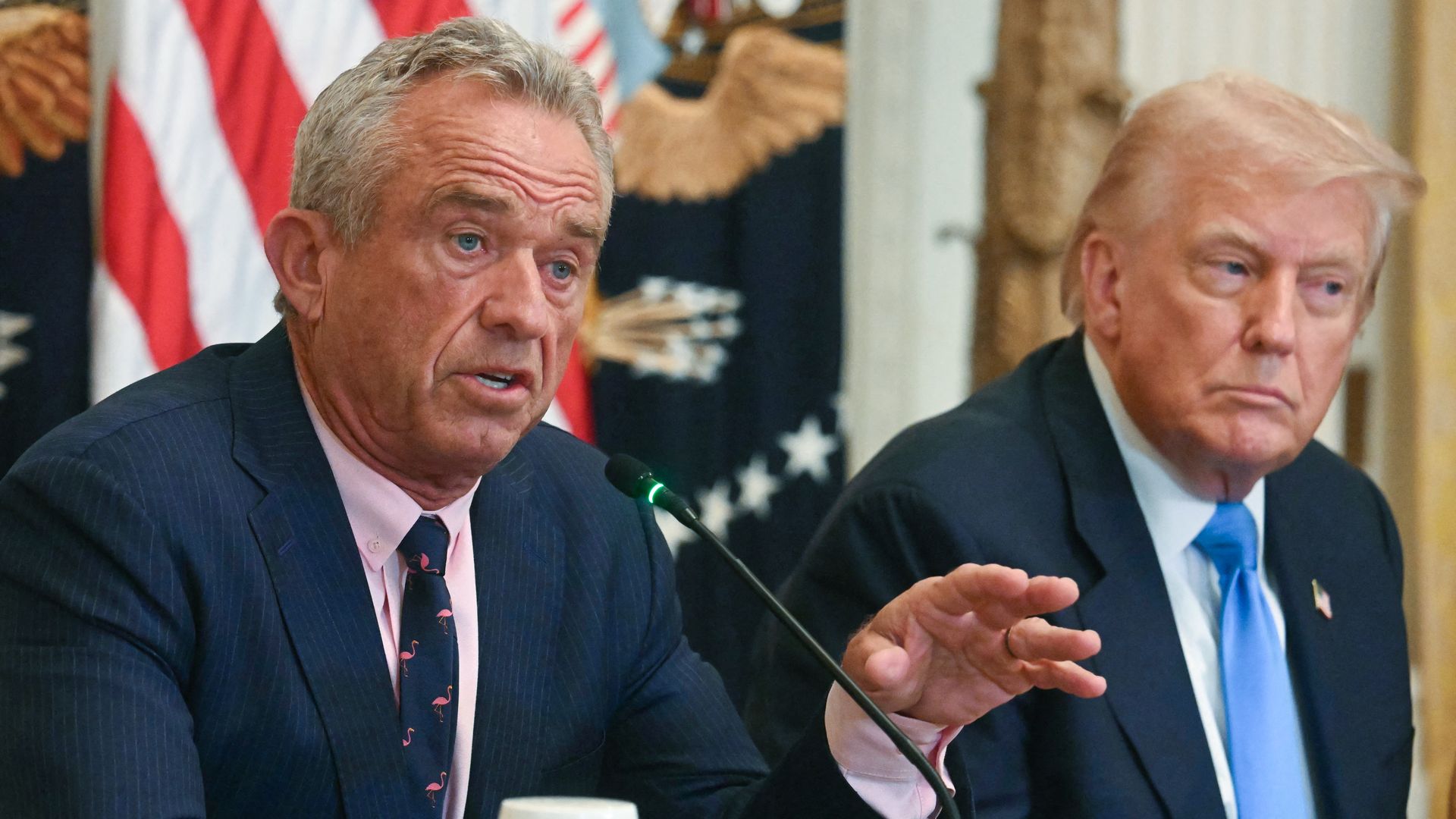 RFK Jr., who is wearing a dark suit, gestures as he speaks into a microphone next to President Trump. An American flag can be seen behind the two men.
