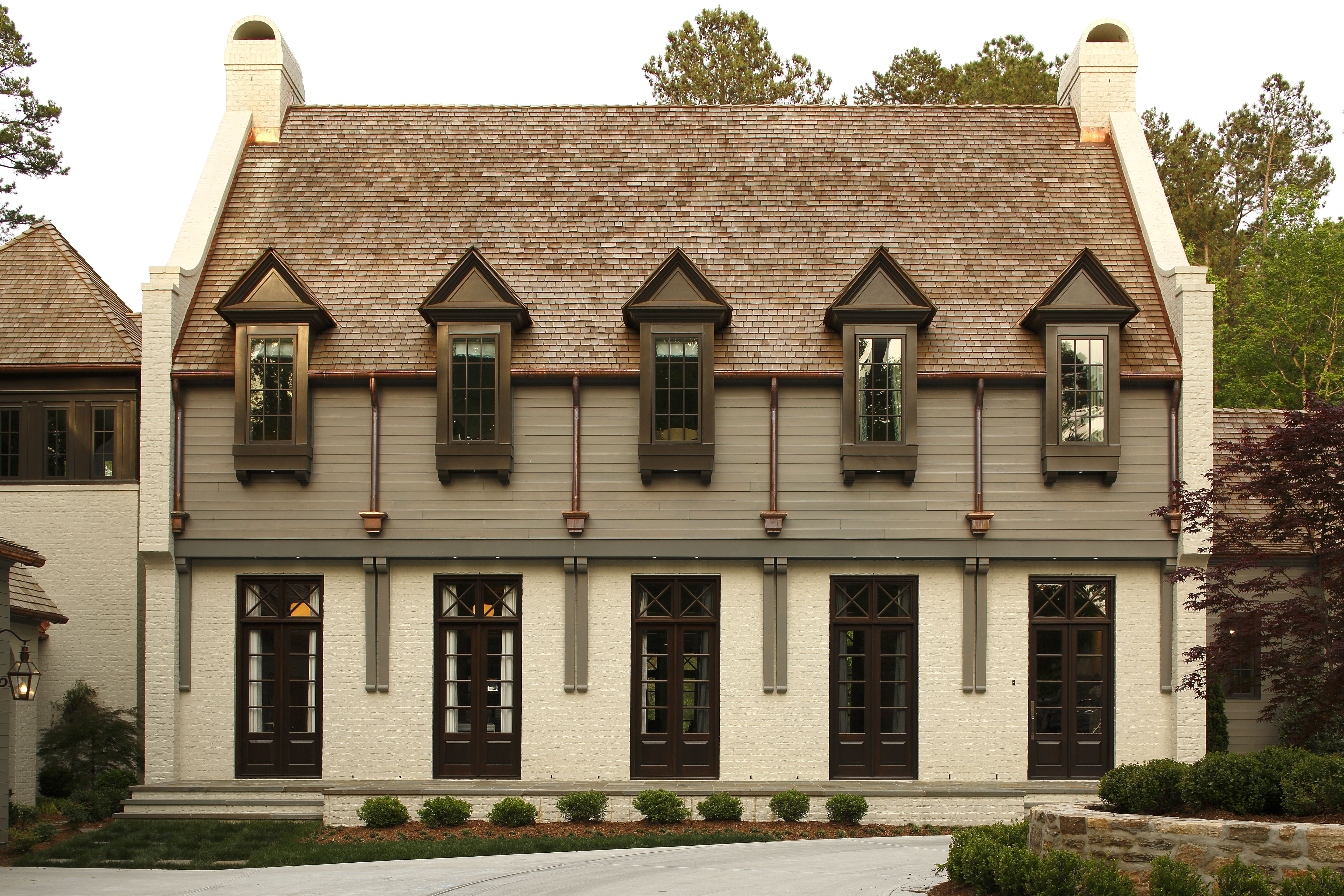 Symmetrical two-story beige house with steep brown shingle roof, five tall dark-framed windows on each floor, white brick walls, and surrounding green shrubs and trees.