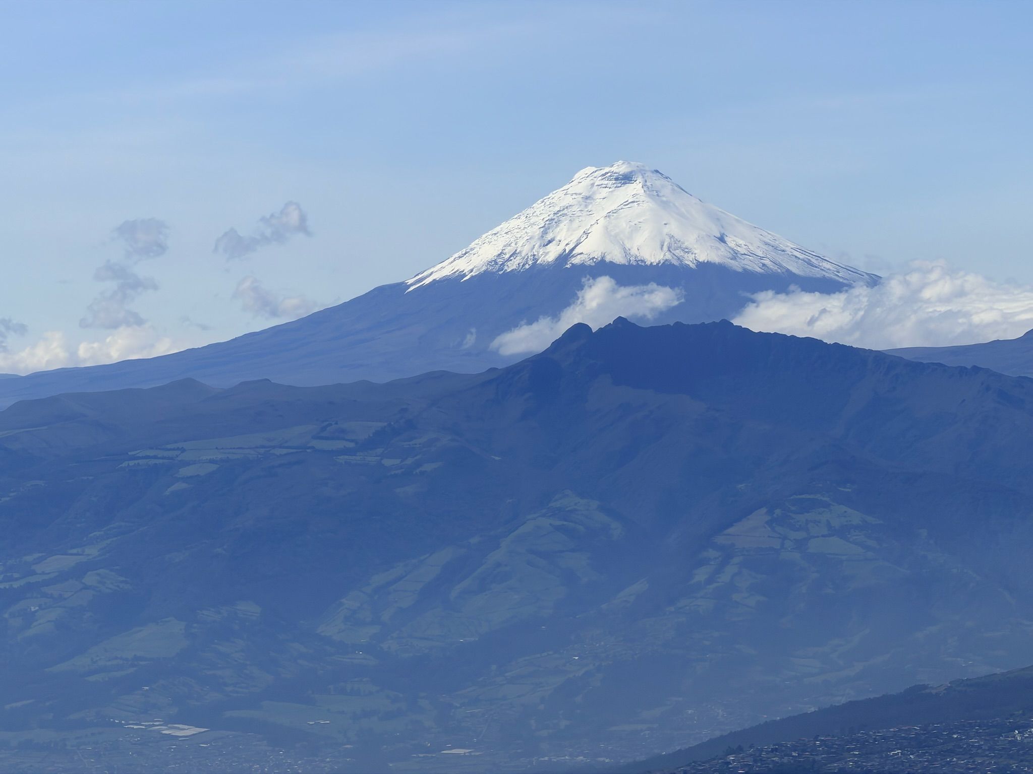 Snow-covered, cone-shaped mountain peak rises above blue ridges and green valleys, under a clear blue sky with scattered clouds.