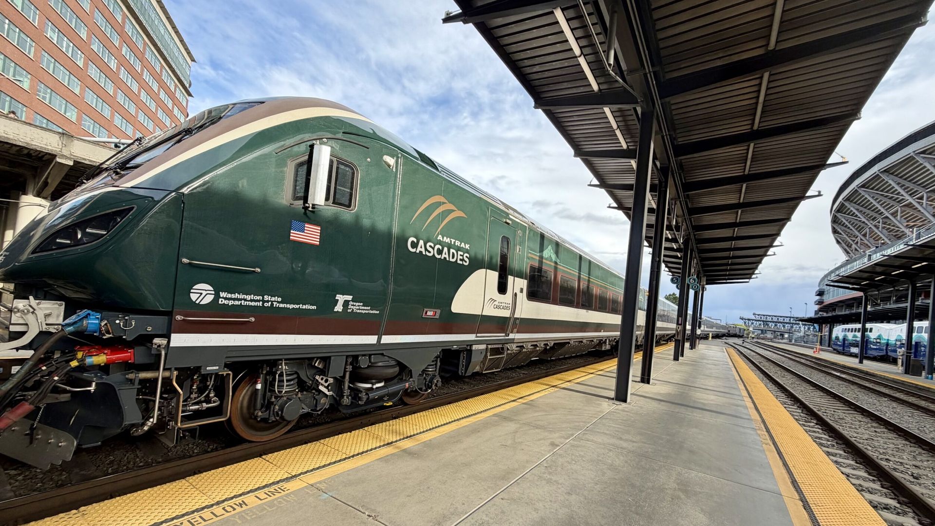 Green Amtrak Cascades locomotive at a station, with a yellow safety edge and tactile paving on the platform. Brick building to the left, a dark metal canopy overhead, and a blue sky.