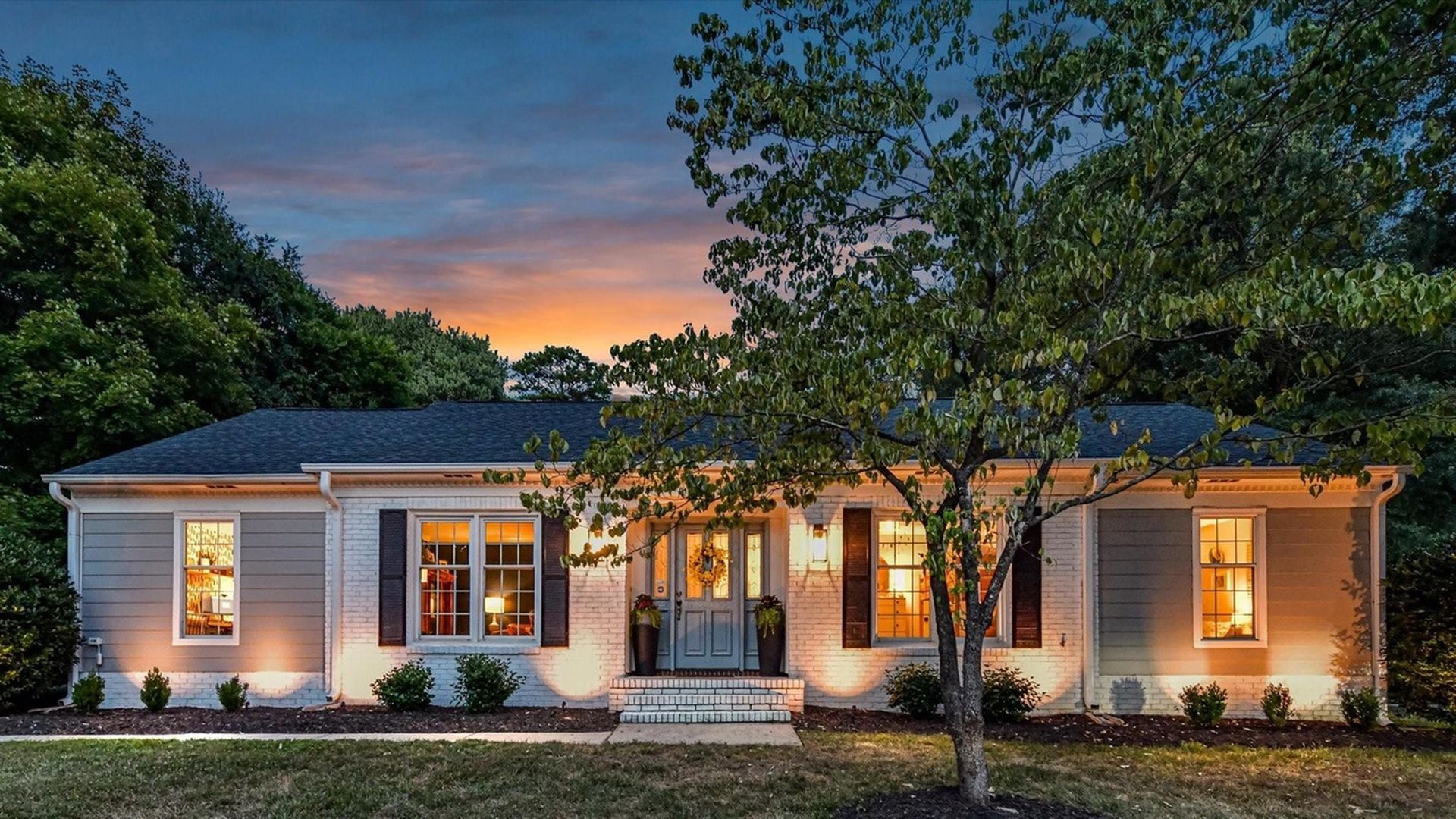 Single-story house with white brick and gray siding, illuminated windows and warm porch light at sunset, tree in front yard, green grass, and dark blue sky.