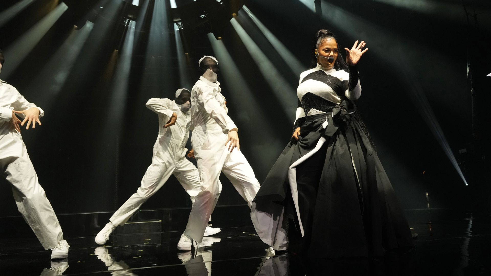 Janet Jackson performing on stage in a black and white outfit with dancers in white behind her