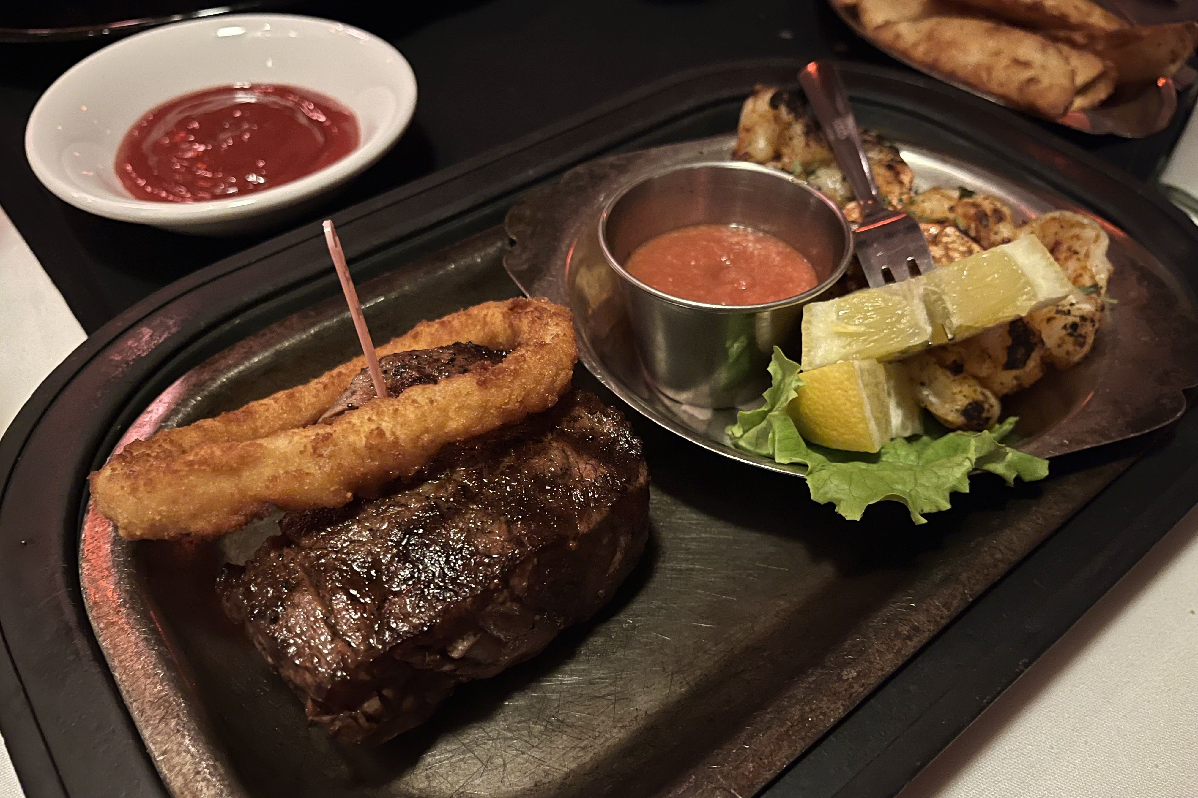 A sizzling steak topped with a crispy onion ring is served on a metal platter. Next to it, there's a side dish with grilled seasoned shrimp, garnished with lemon wedges and lettuce. 