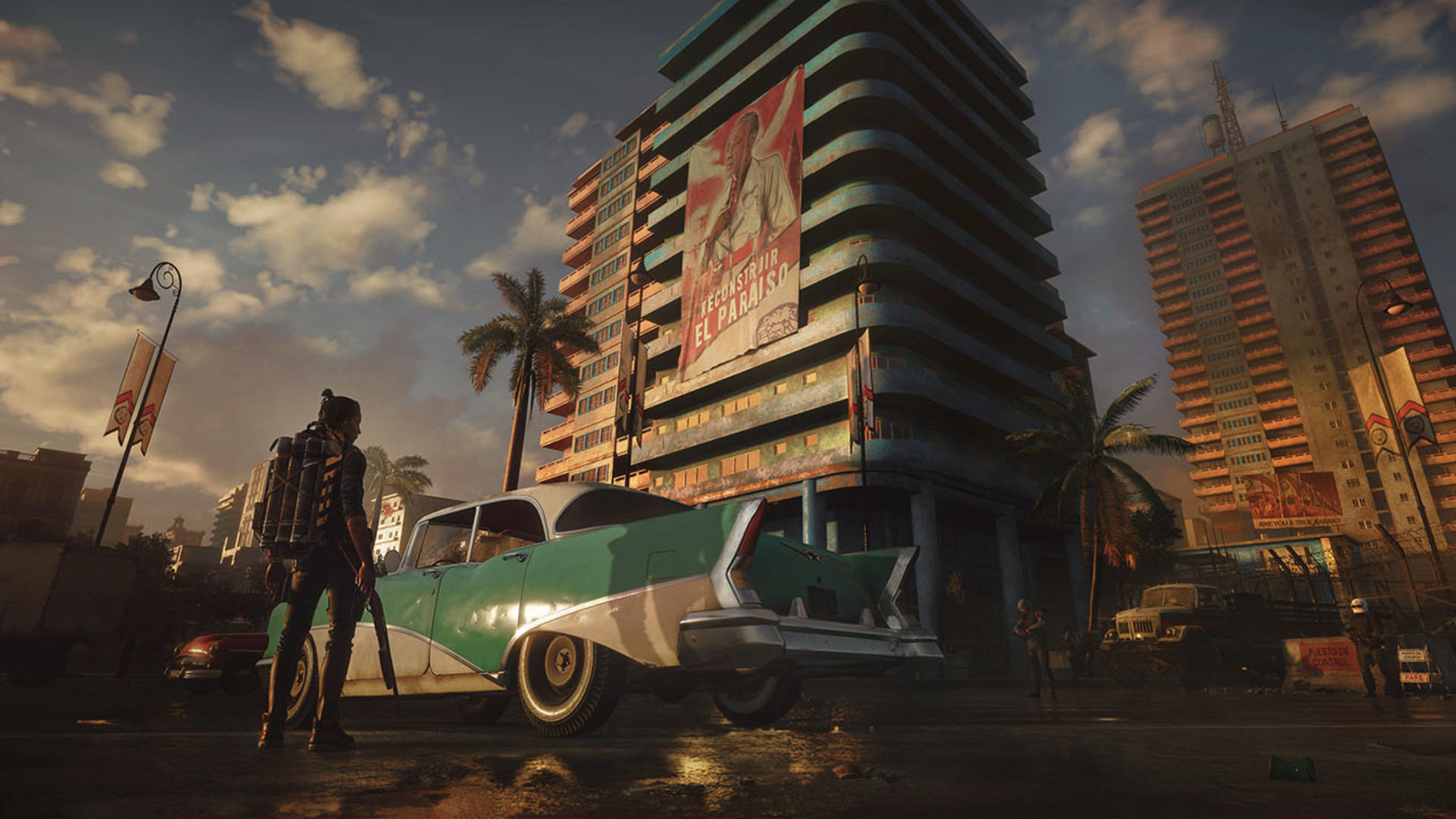 Graphic image of a scene from Far Cry 6, with a character standing in front of a vintage car and towering building