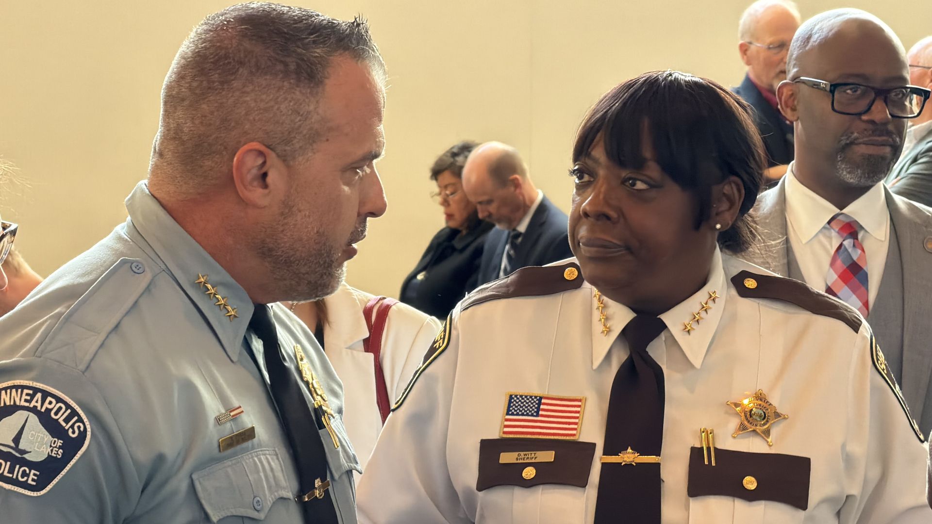 Two police officers converse in a crowded room: a light-blue uniformed man on the left and a Black woman in a white uniform with gold badges and an American flag patch, others blurred in background.