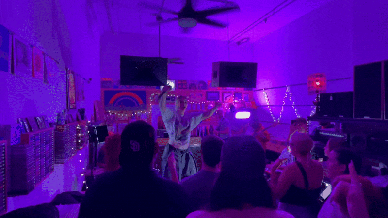 A performer sings into a microphone at an intimate concert with purple lighting. The small venue has a cozy crowd, vinyl records on the wall, fairy lights, and a neon “Speck’s Records & Tapes” sign.