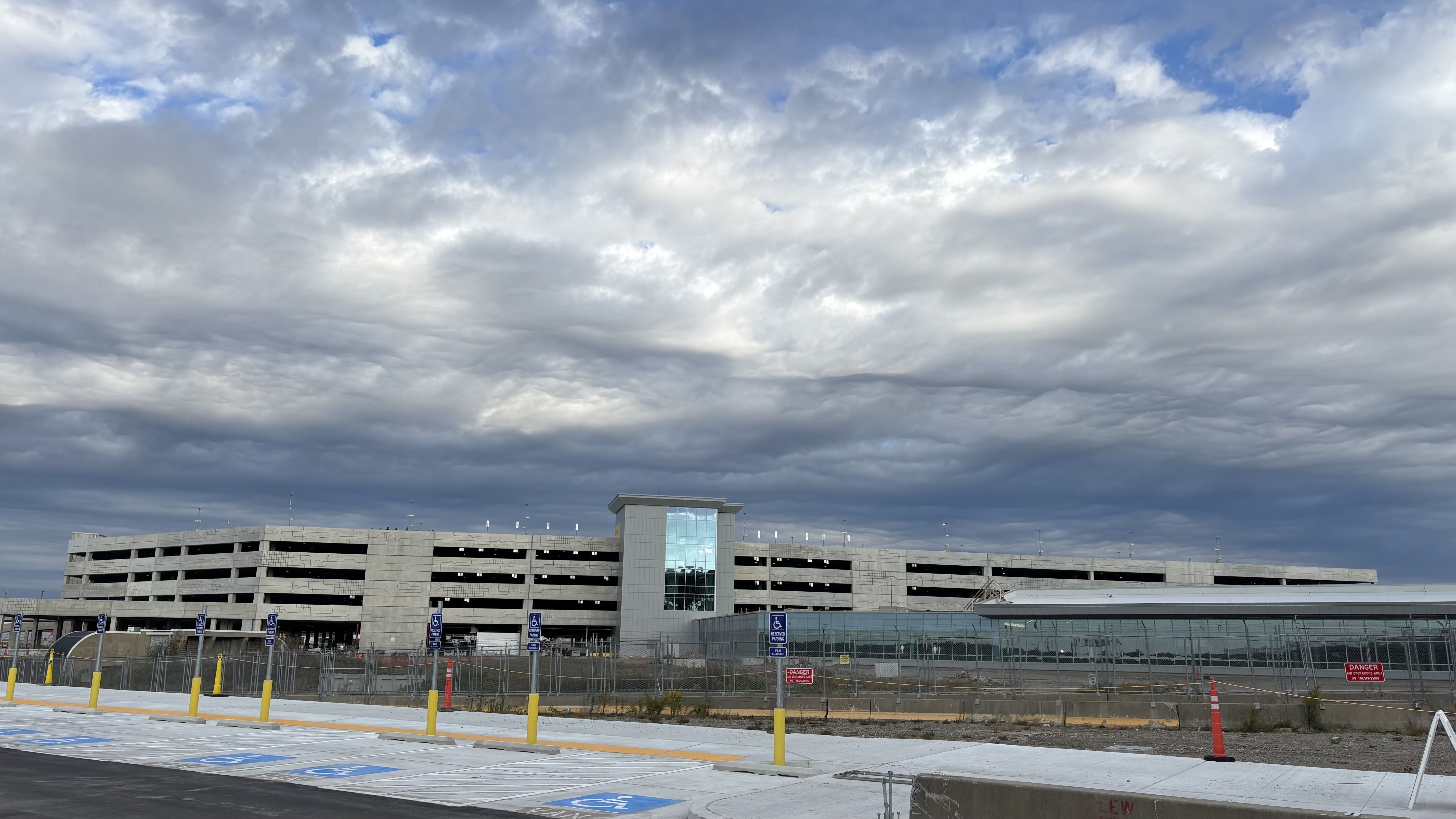 Large gray multi-story parking garage with a central glass elevator shaft under a dramatic cloudy sky. Empty handicap parking spaces and yellow posts in foreground.