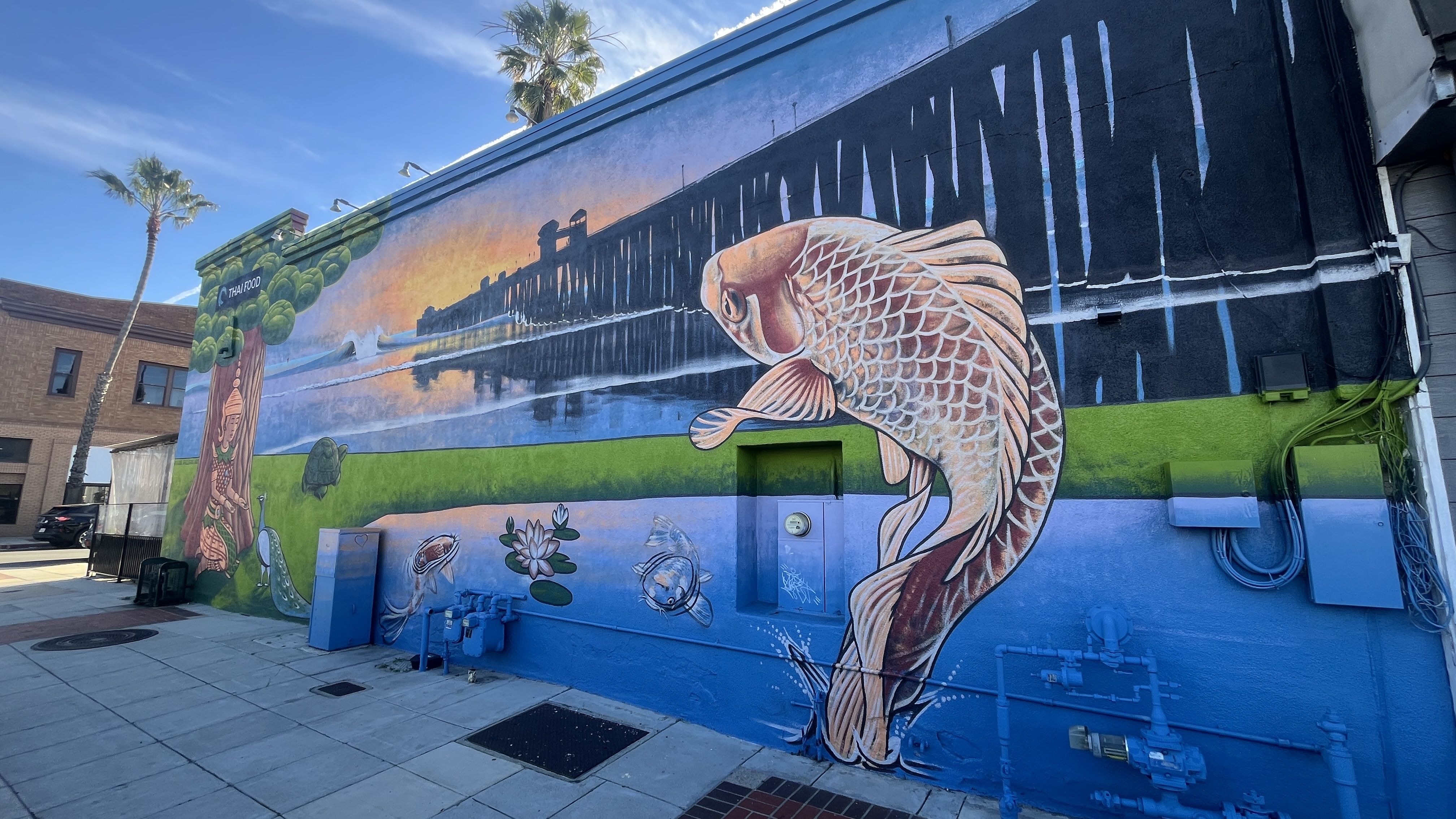 Vibrant mural on a building wall features a large orange koi with white scales leaping out of a pond and looking the Oceanside pier at sunset. 