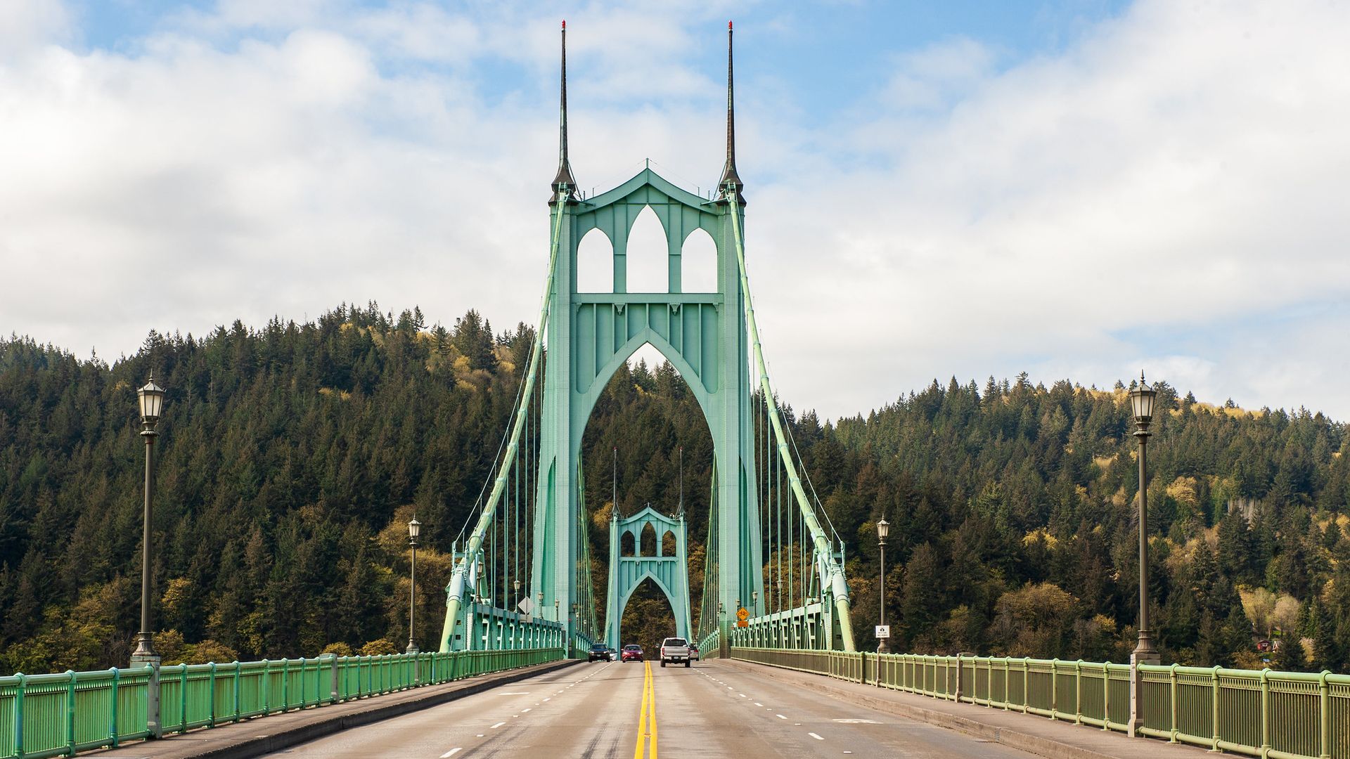 Teal-green suspension bridge with tall towers and arched openings, crossing a road through a forested hillside under a blue sky with clouds.