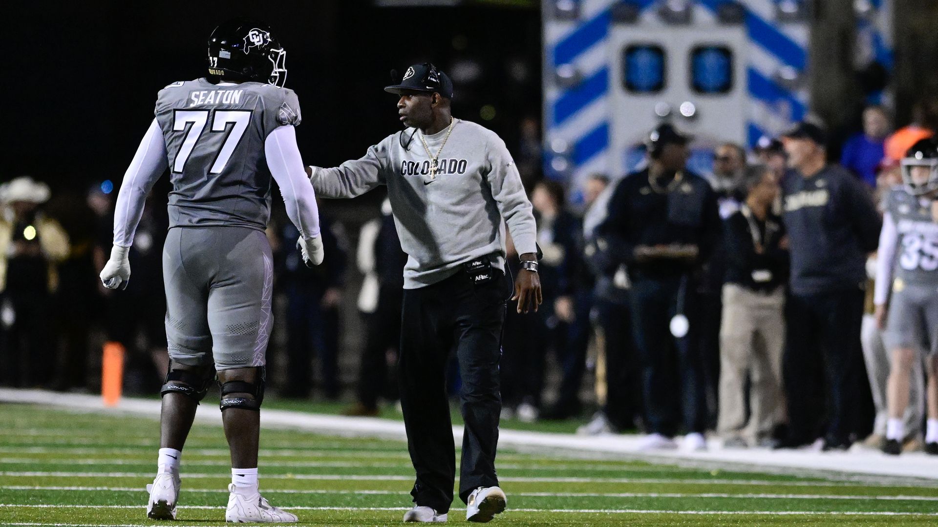 Deion Sanders in gray Colorado sweatshirt talking to Jordan Seaton in gray uniform with black helmet on green field at night.