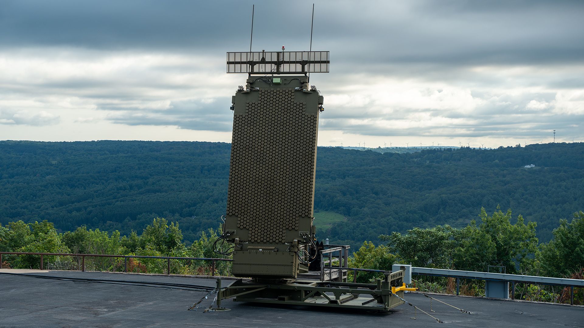 Large military radar unit with hexagonal panel pattern on platform outdoors, green hills and cloudy sky in background.