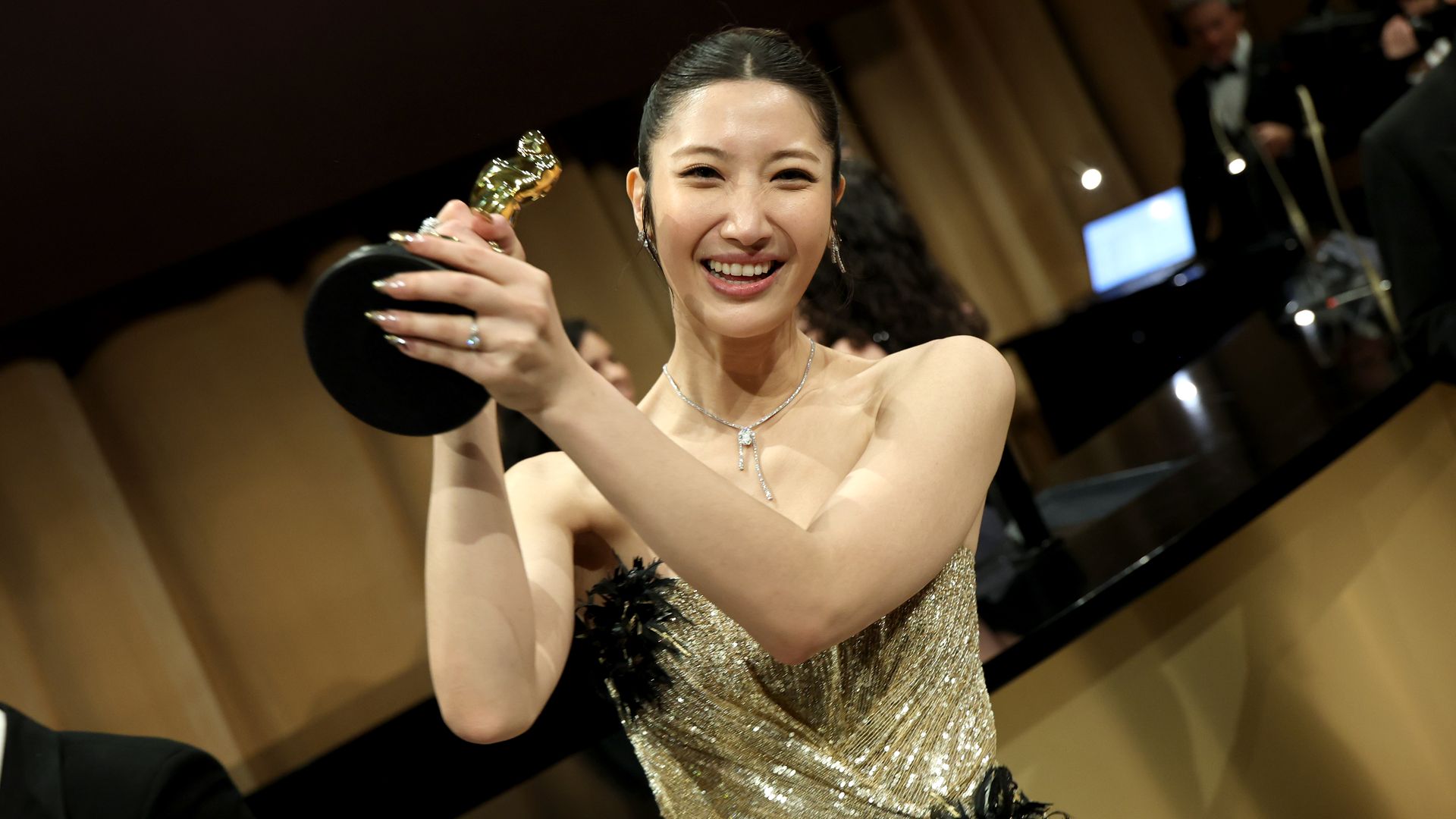 Smiling woman in a sparkling gold evening gown lifts a small gold trophy on a stage at a formal awards ceremony, wearing a silver necklace and earrings.