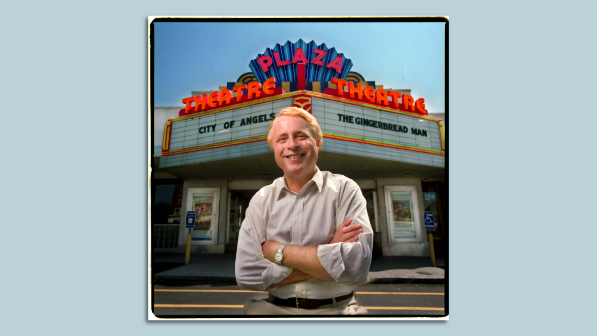 George LeFont stands smiling and with him arms cross outside the Plaza Theatre neon marquee