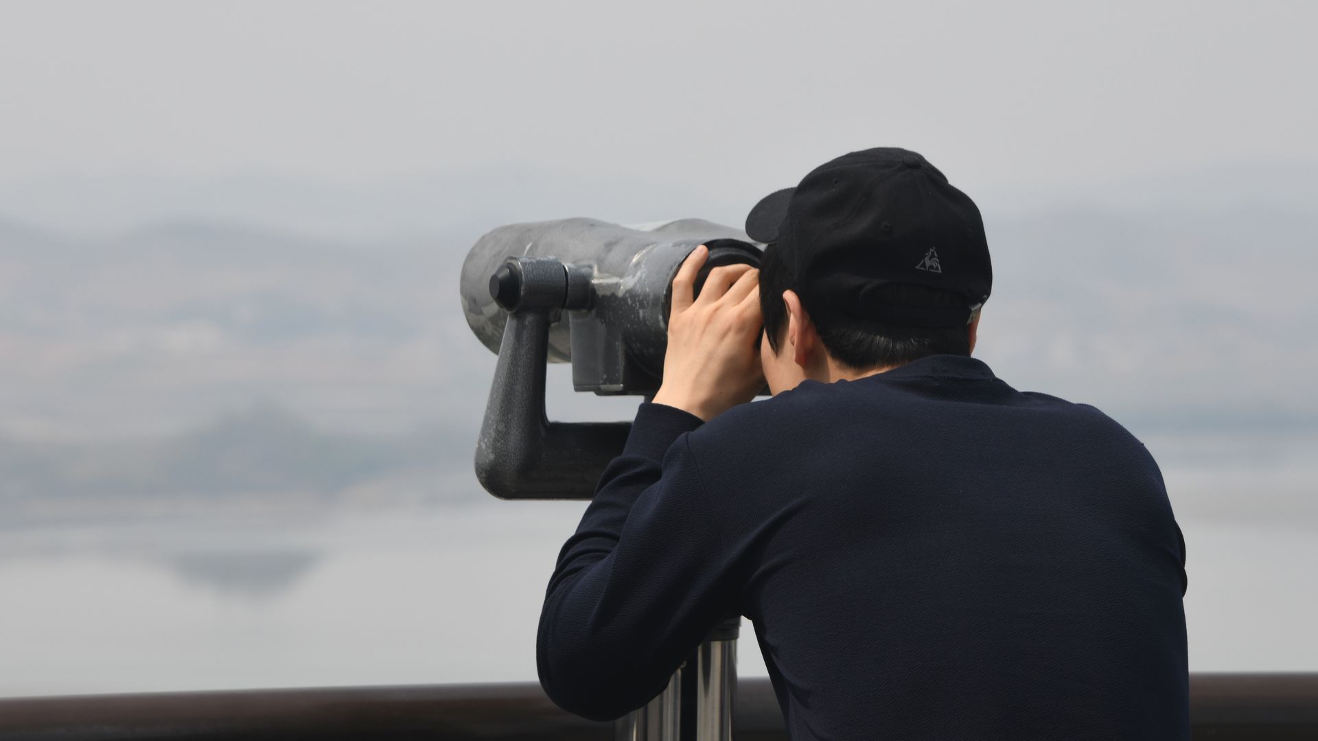 A visitor, wearing all black, looks towards North Korea through binoculars at a South Korean observatory, with a white sky.