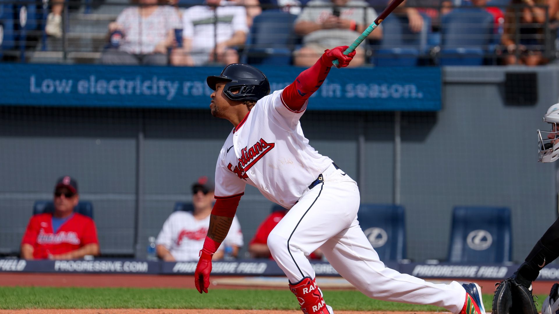 A baseball player in a white and red Indians jersey swings a teal-taped bat, wearing a black helmet, as fans watch from blurred stands in the background.