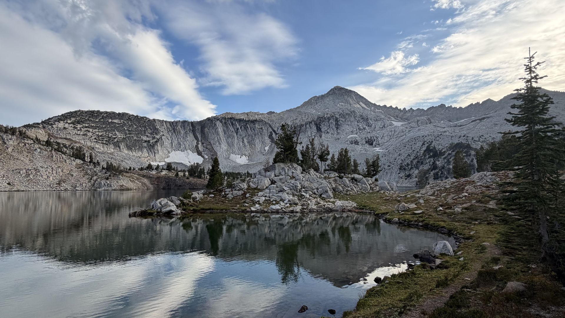 Rocky, snow-dusted mountains reflect in a calm alpine lake; foreground shows a rocky shore with evergreens, under a blue sky with wispy white clouds.