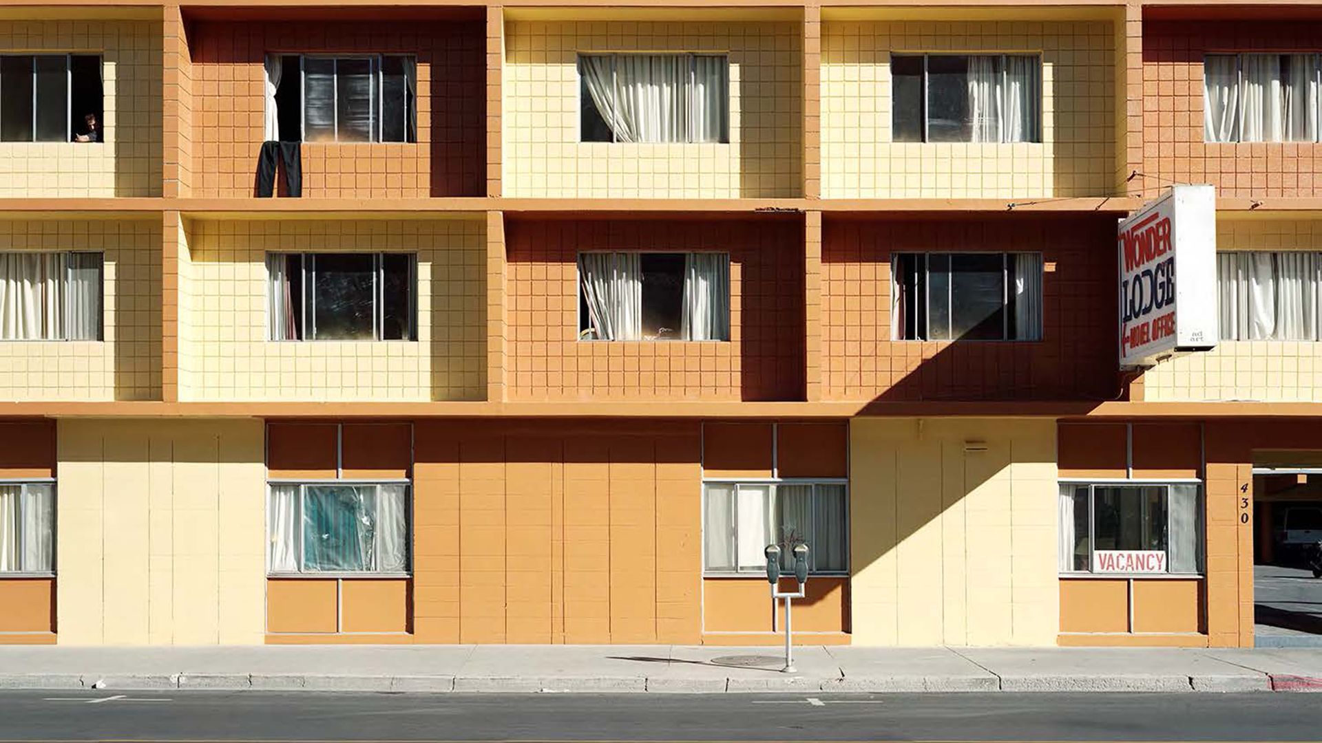 Beige and orange tiled apartment building with square balconies and windows. Two people lean on a balcony near the left-center while a bright blue sky lies above; a vacancy sign is on the ground to the right.