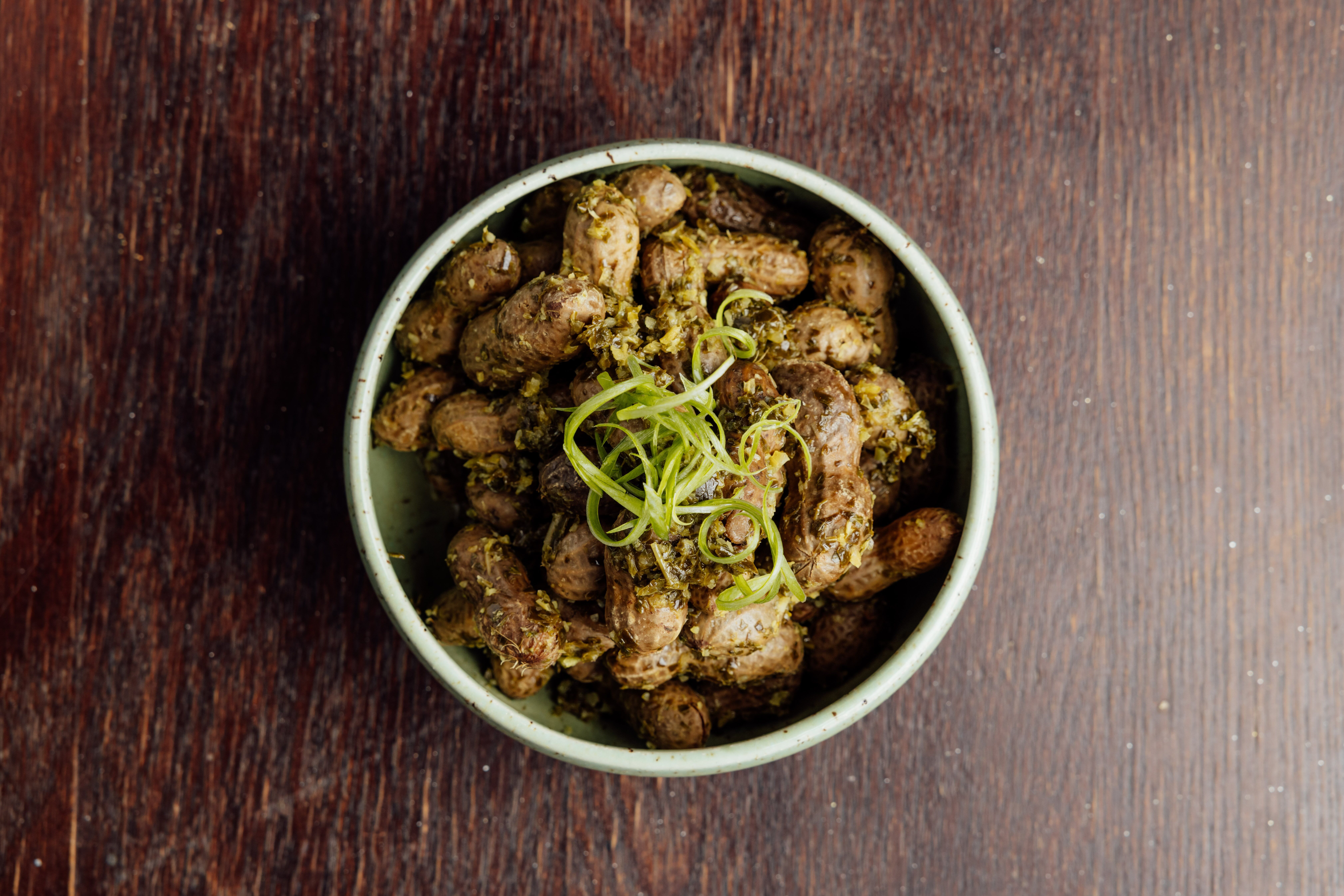 Top-down view of a light green ceramic bowl filled with browned, sauteed meat bites, garnished with chopped herbs and curly green onion, set on a dark wooden table.