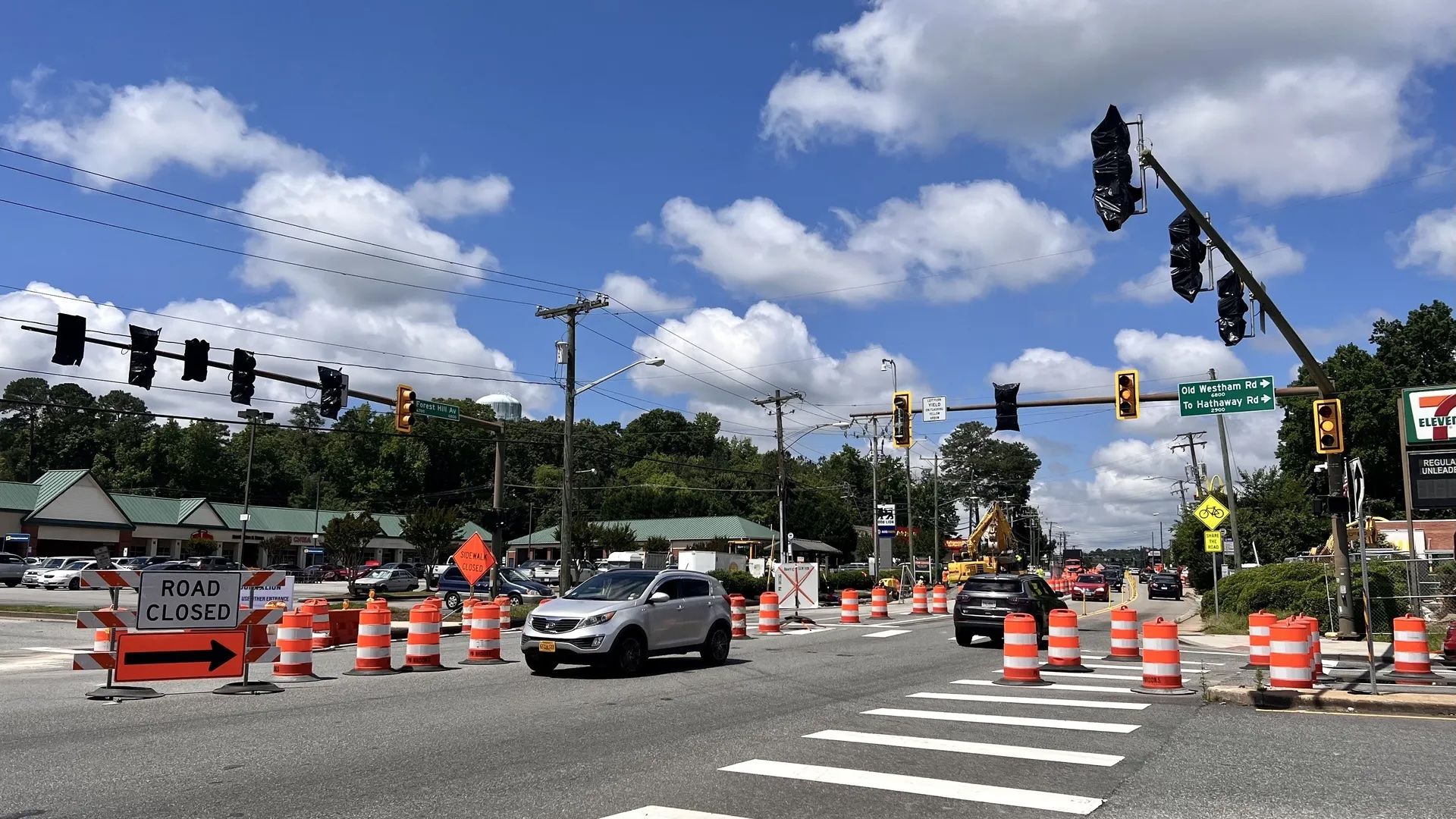 Busy intersection under construction with orange cones and a ROAD CLOSED sign. Blacked-out traffic lights, crosswalk lines, several cars, green signs, and a bright blue sky.