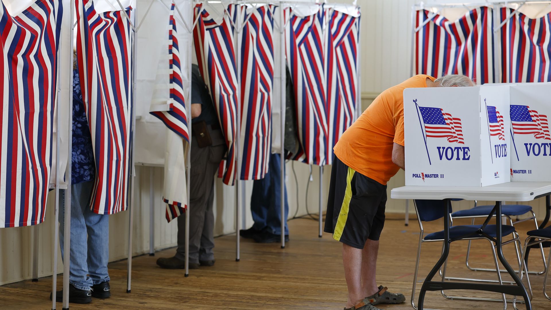 Indoor polling station with red, white, and blue striped voting booths. A person in an orange shirt stands at a booth labeled "VOTE" with American flag graphics; wooden floor and chairs nearby.