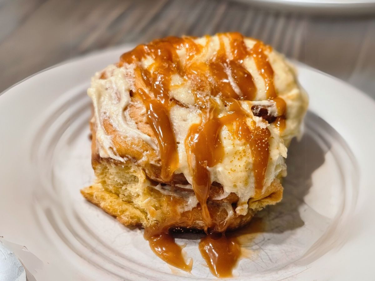 A cinnamon bun topped with tan caramel sauce on a white ceramic plate.